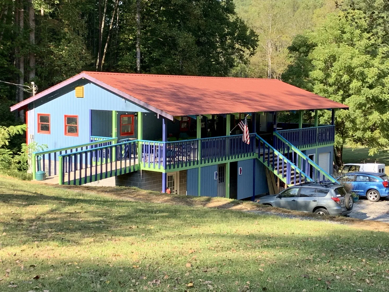 Blue house with red roof and purple porch on a grassy hillside, surrounded by trees