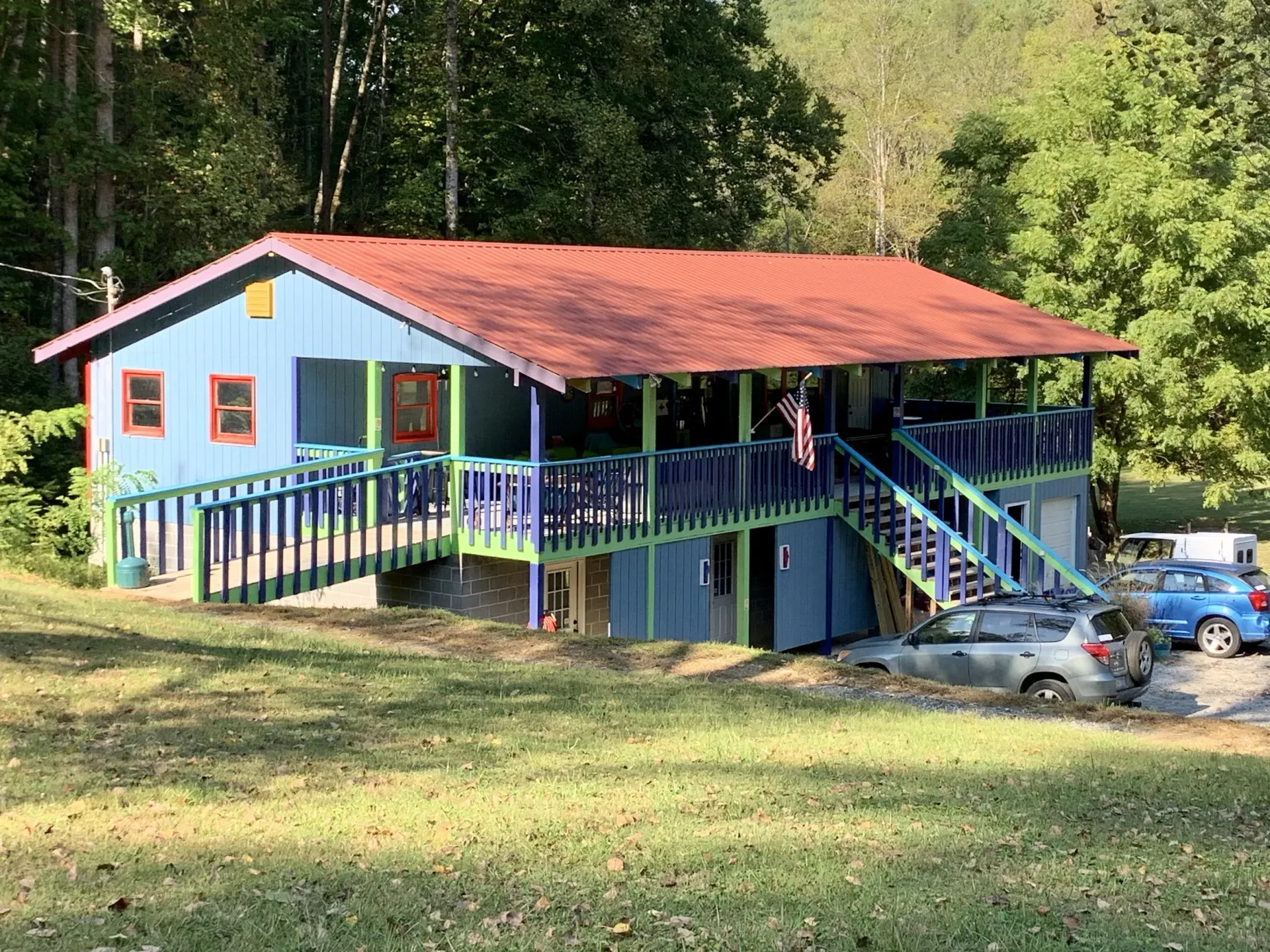 Blue cabin with red roof and porch stairs in a grassy wooded area, with cars parked nearby