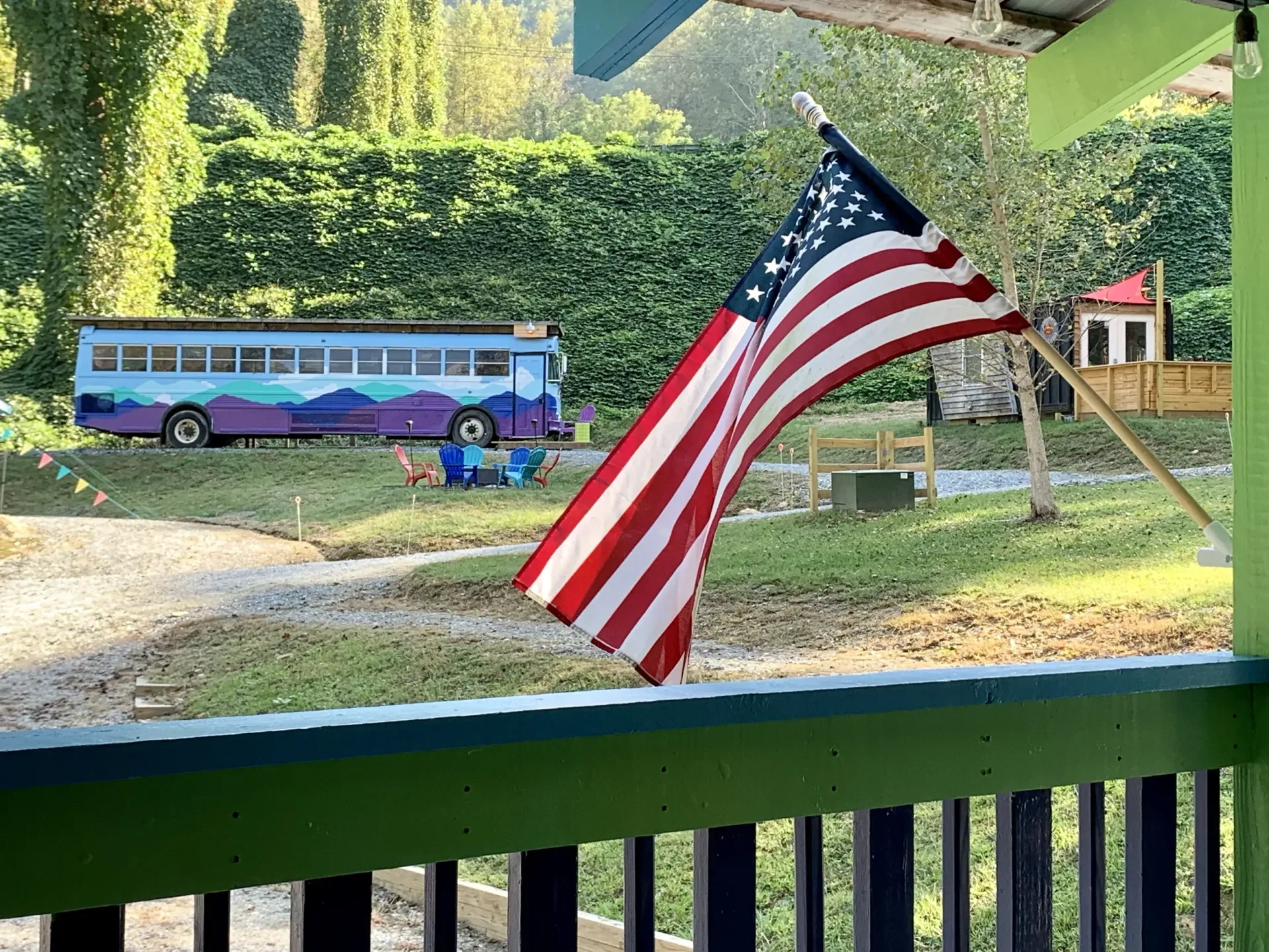American flag hanging in front of a hedge, with a colorful school bus and trees in the background.