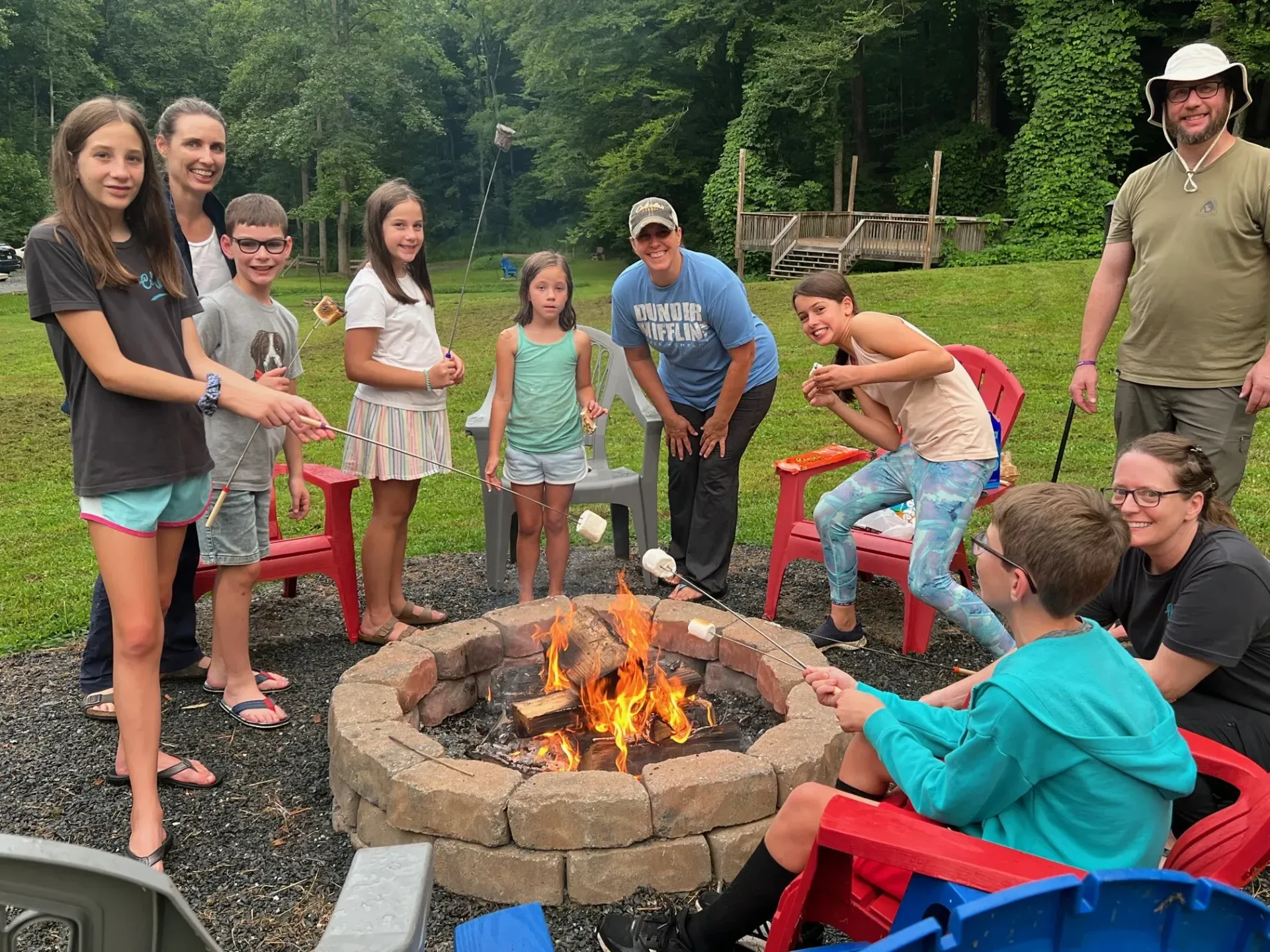 Group gathered around a fire pit outdoors, smiling and roasting marshmallows on red chairs.