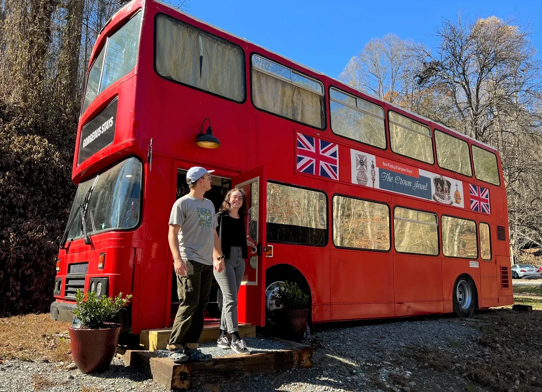 Red double-decker bus with a Union Jack side, parked outdoors; two people stand at the front door.