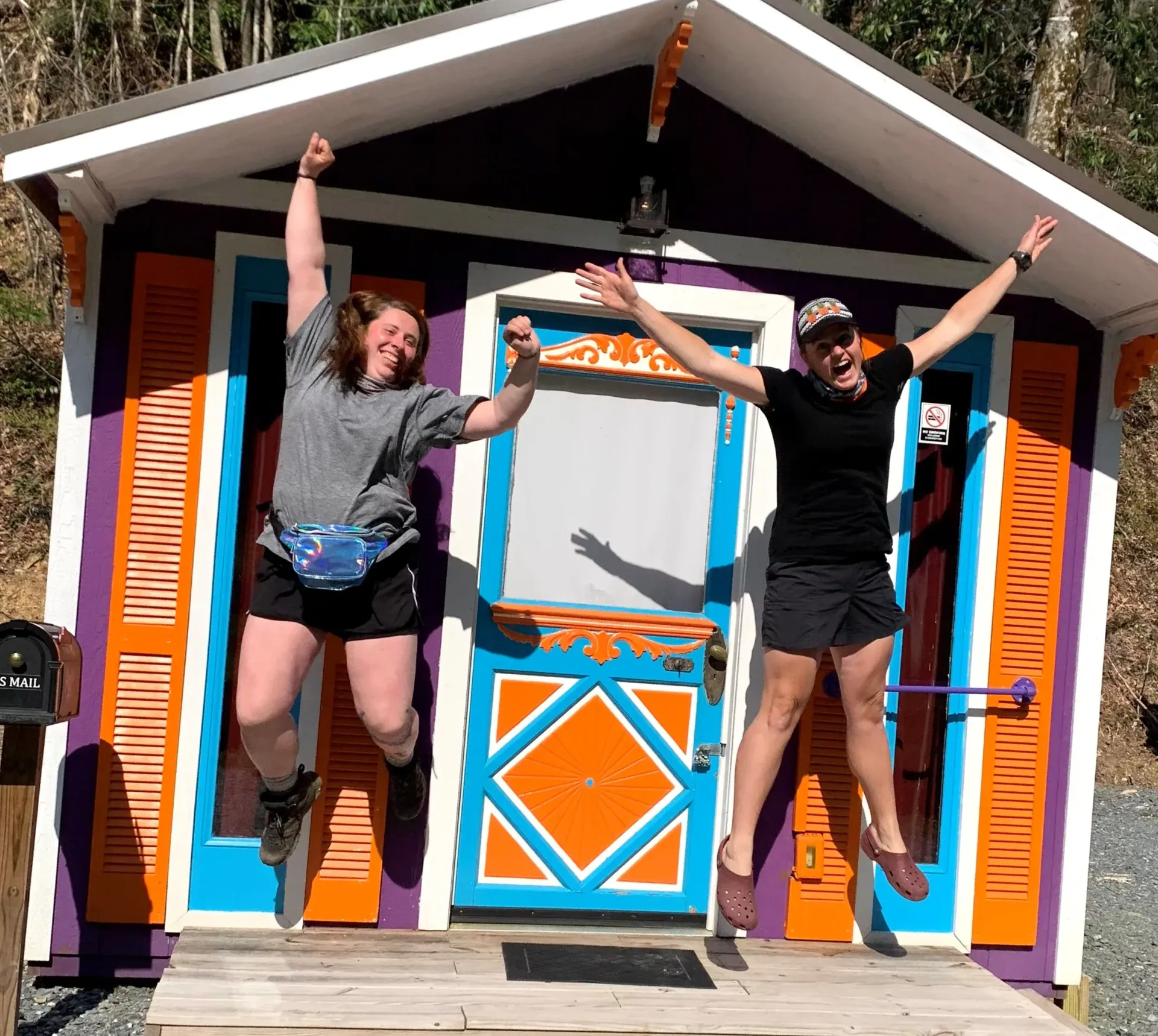 Two people jumping in front of a colorful cabin with orange and blue trim