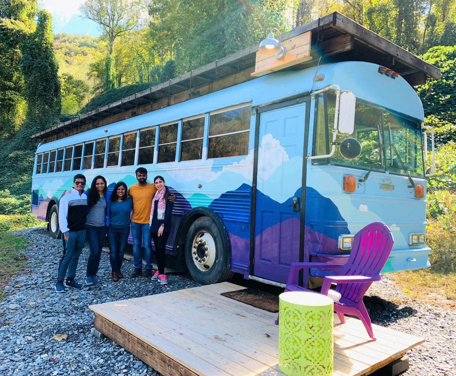 Five people stand beside a blue-purple bus parked on a gravel road in a wooded area.