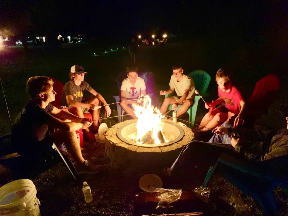 Friends sit around a glowing campfire at night, gathered in folding chairs outdoors.