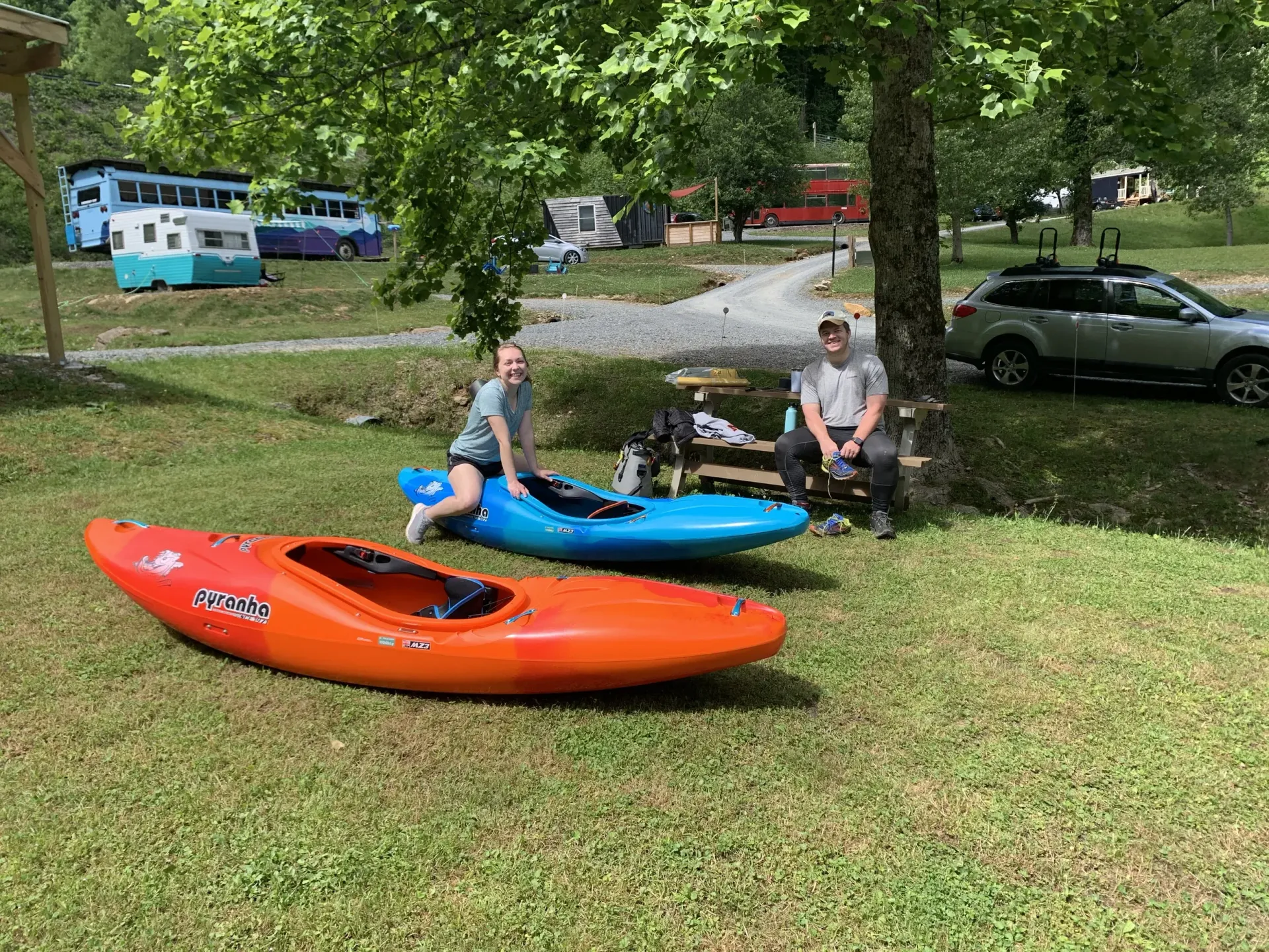 Two people with kayaks on a grassy park hill near parked cars and trees