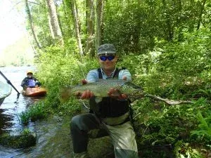 Two kayakers in a narrow forest creek, one holding a fish in the foreground.