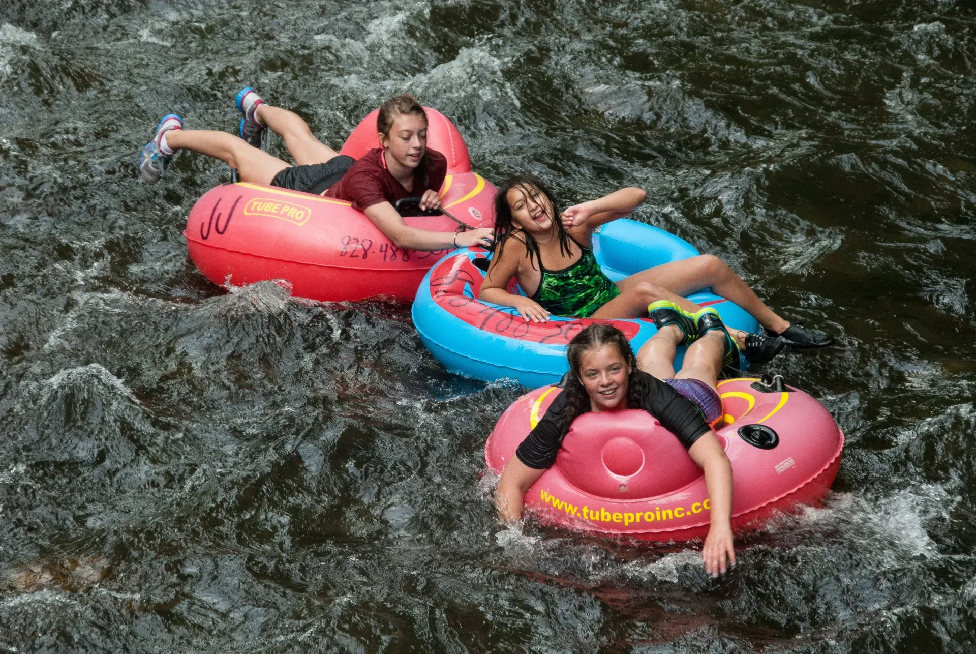 Three people tubing down a fast river in colorful inner tubes, splashing through whitewater.