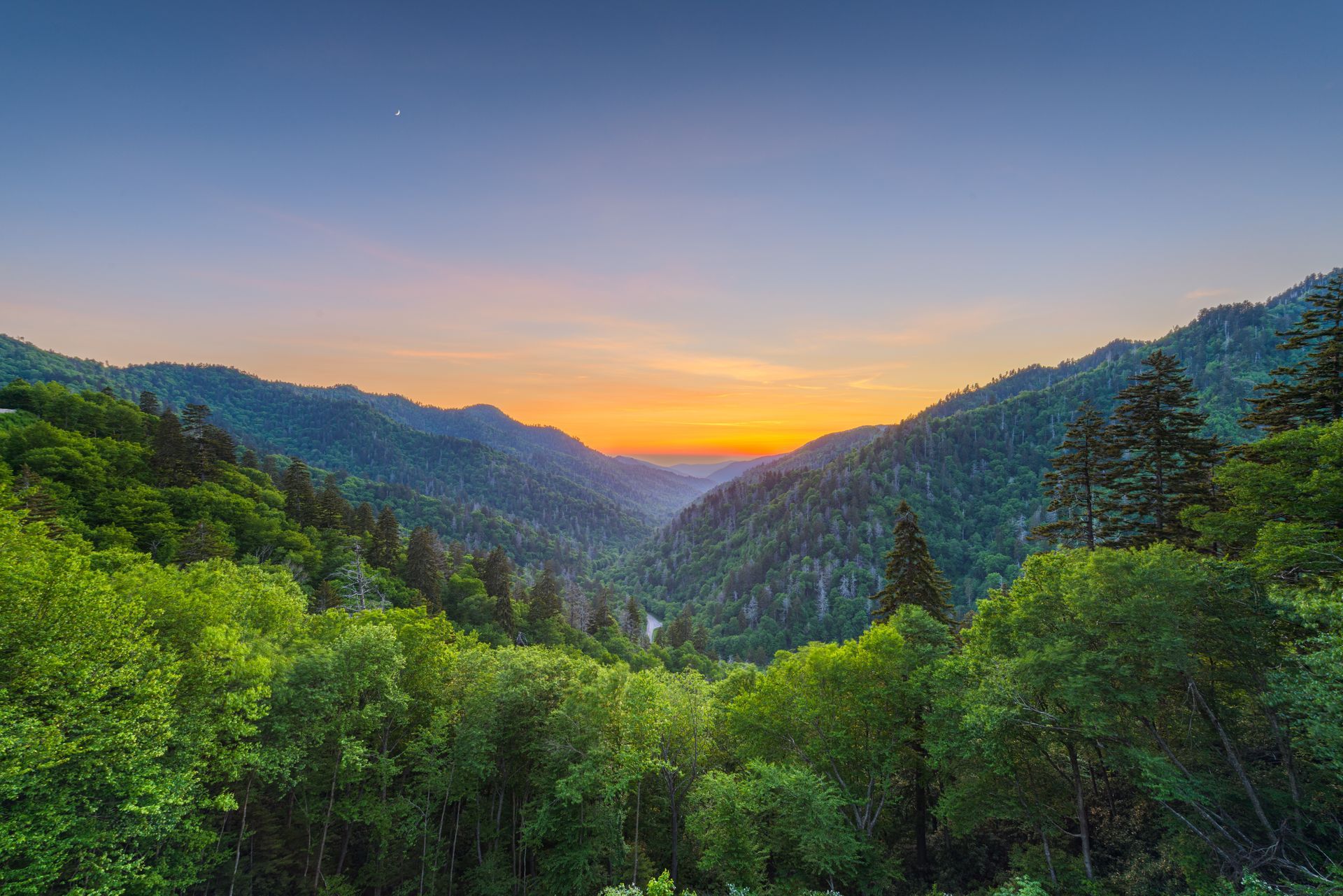Sunrise over a lush green mountain valley with a glowing orange horizon and blue sky