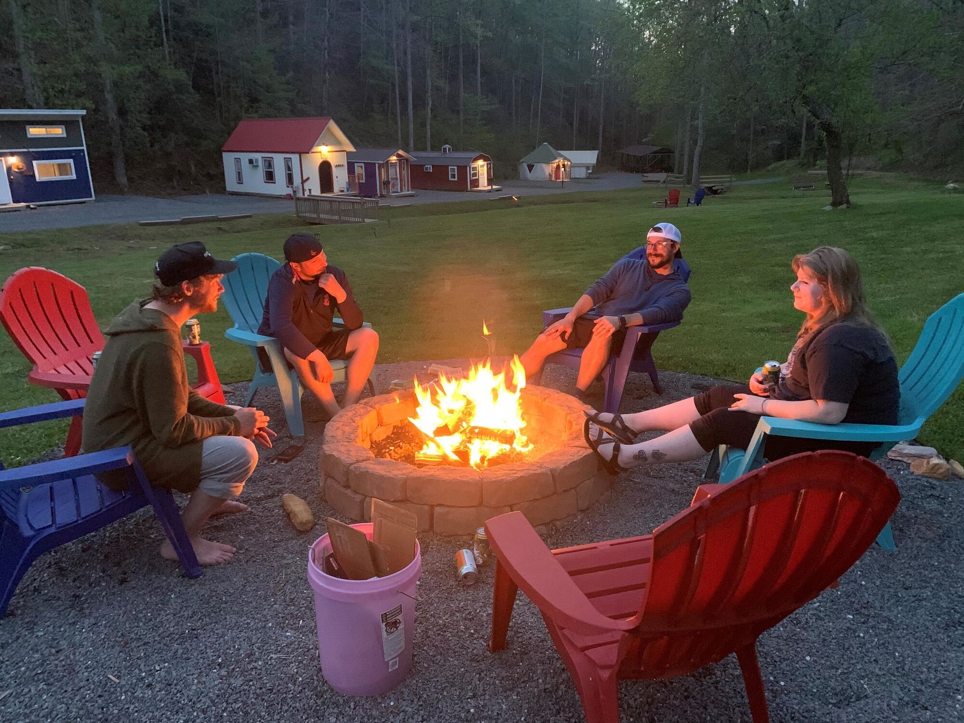 People sitting around a fire pit at dusk in a backyard, with colorful chairs and a house in the background