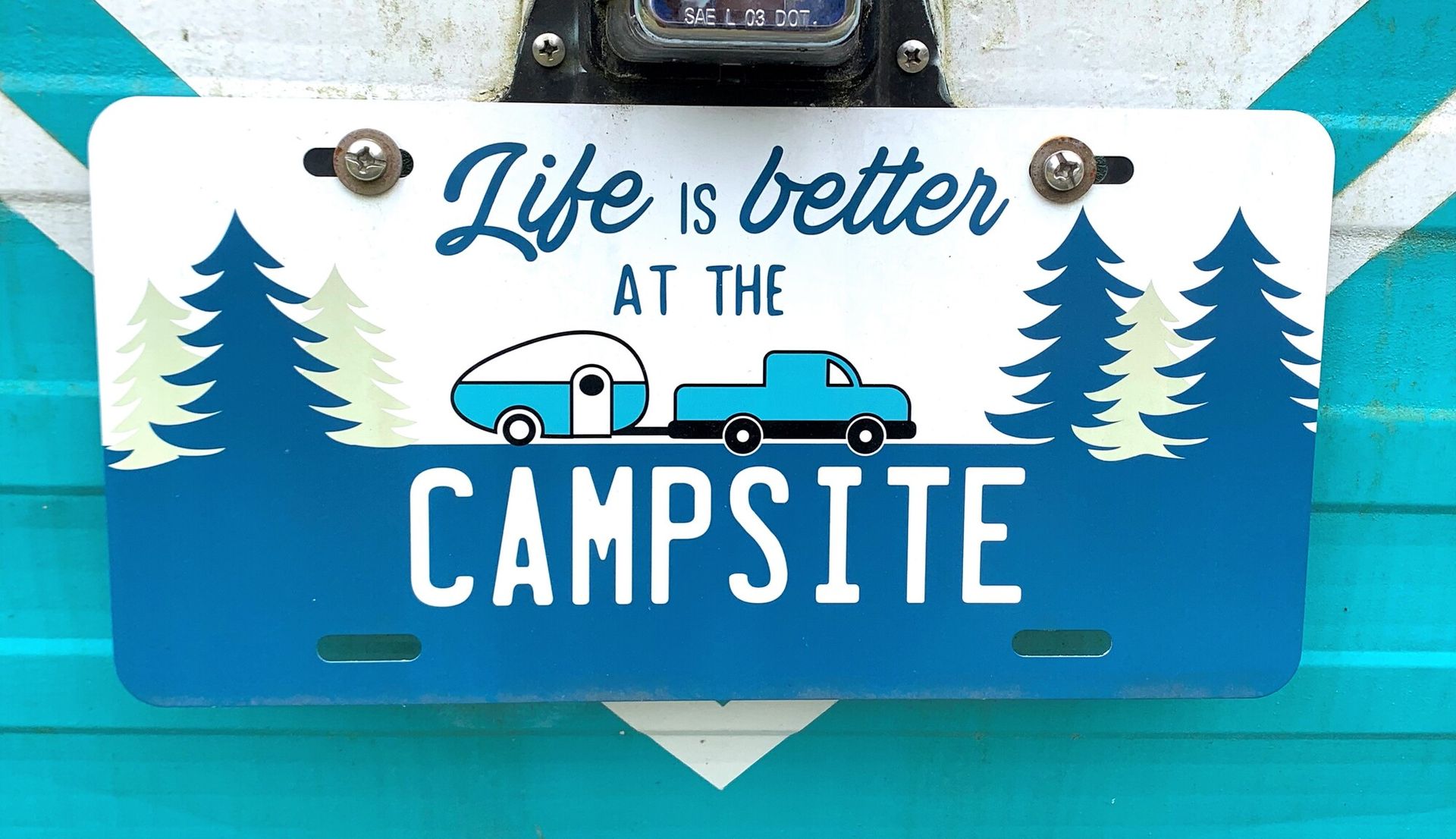 Decorative sign reading “Life is better at the campsite” with a camper trailer, truck, and pine trees on blue background