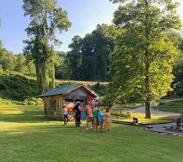 Children gathered by a small wooden shed in a sunny green park with tall trees and a pond