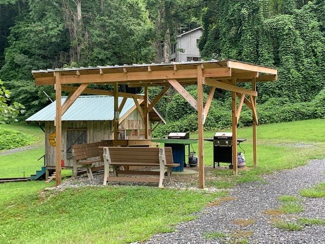 Wooden pavilion with picnic table and grill in a grassy clearing beside a gravel path and forest.