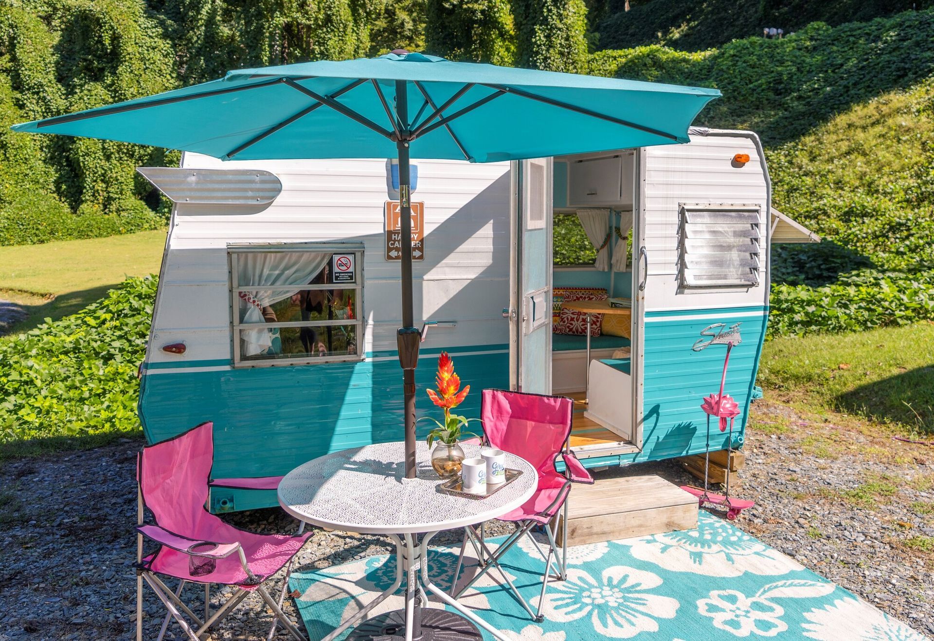 Teal-and-white camper with patio set, pink chairs, umbrella, and floral rug in a sunny wooded campsite