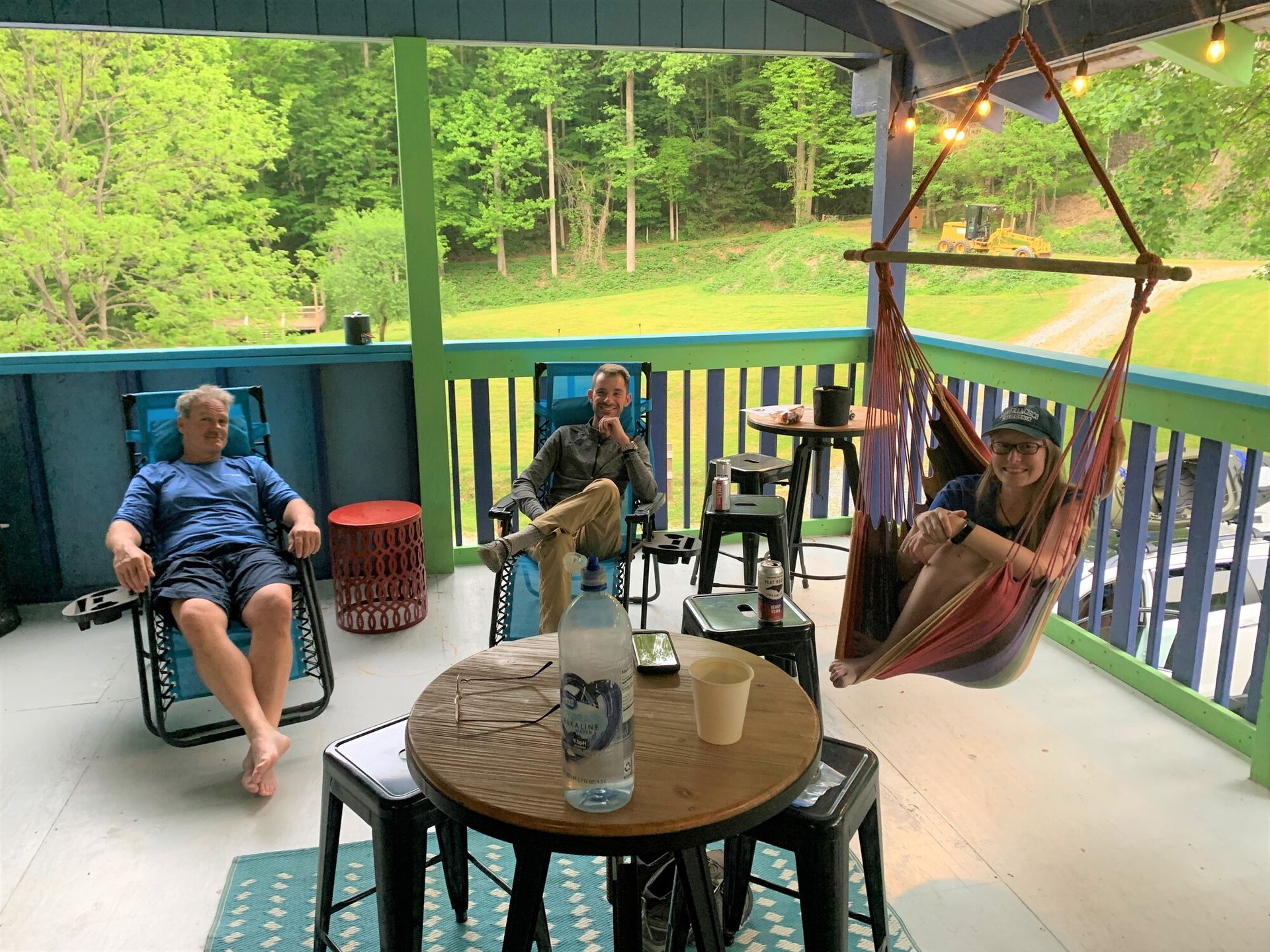 People relaxing on a covered porch with chairs, a table, and a green wooded view beyond the railing.