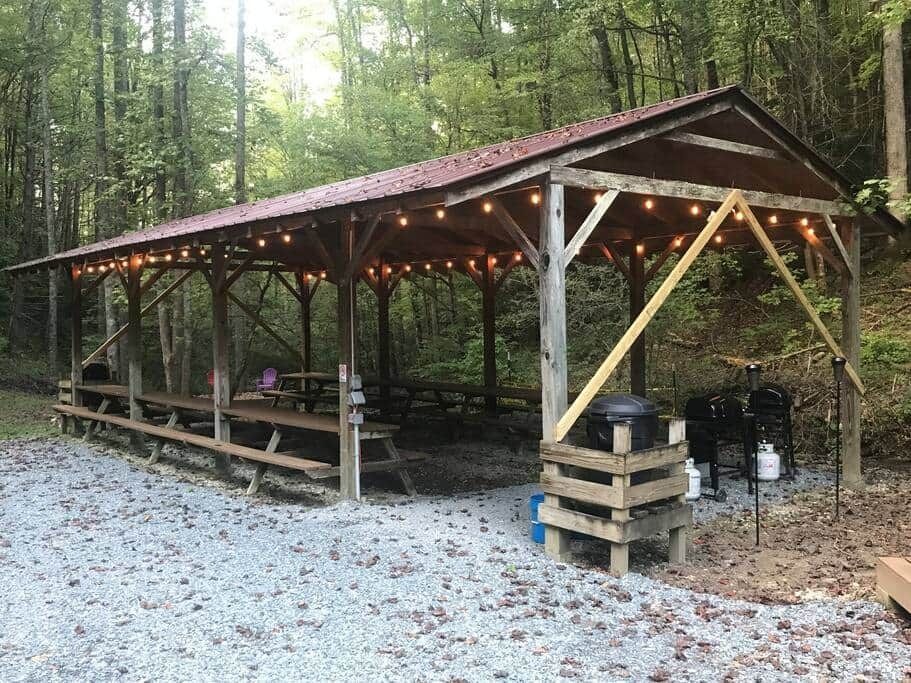 Open-sided wooden pavilion with string lights in a forest clearing, beside a gravel area