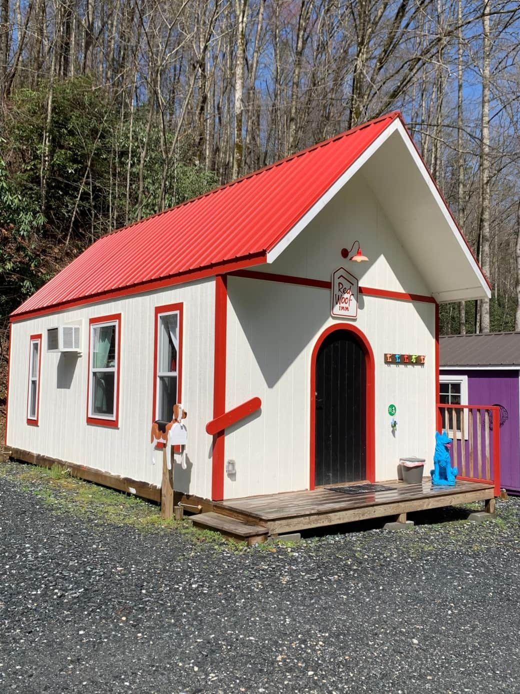 Small white cottage with a red roof and trim on a gravel lot beside trees