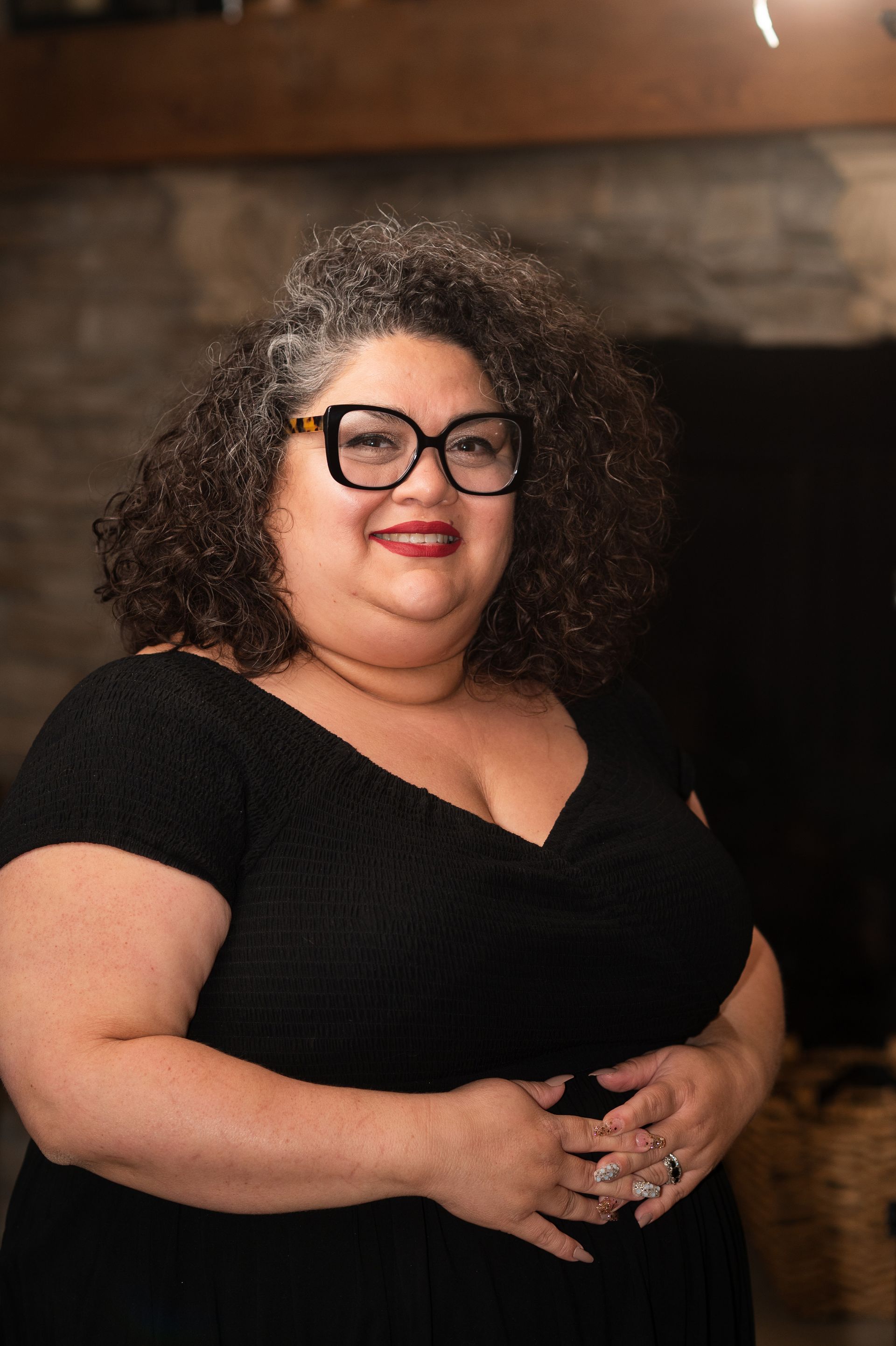 A woman wearing glasses and a black dress is standing in front of a fireplace.