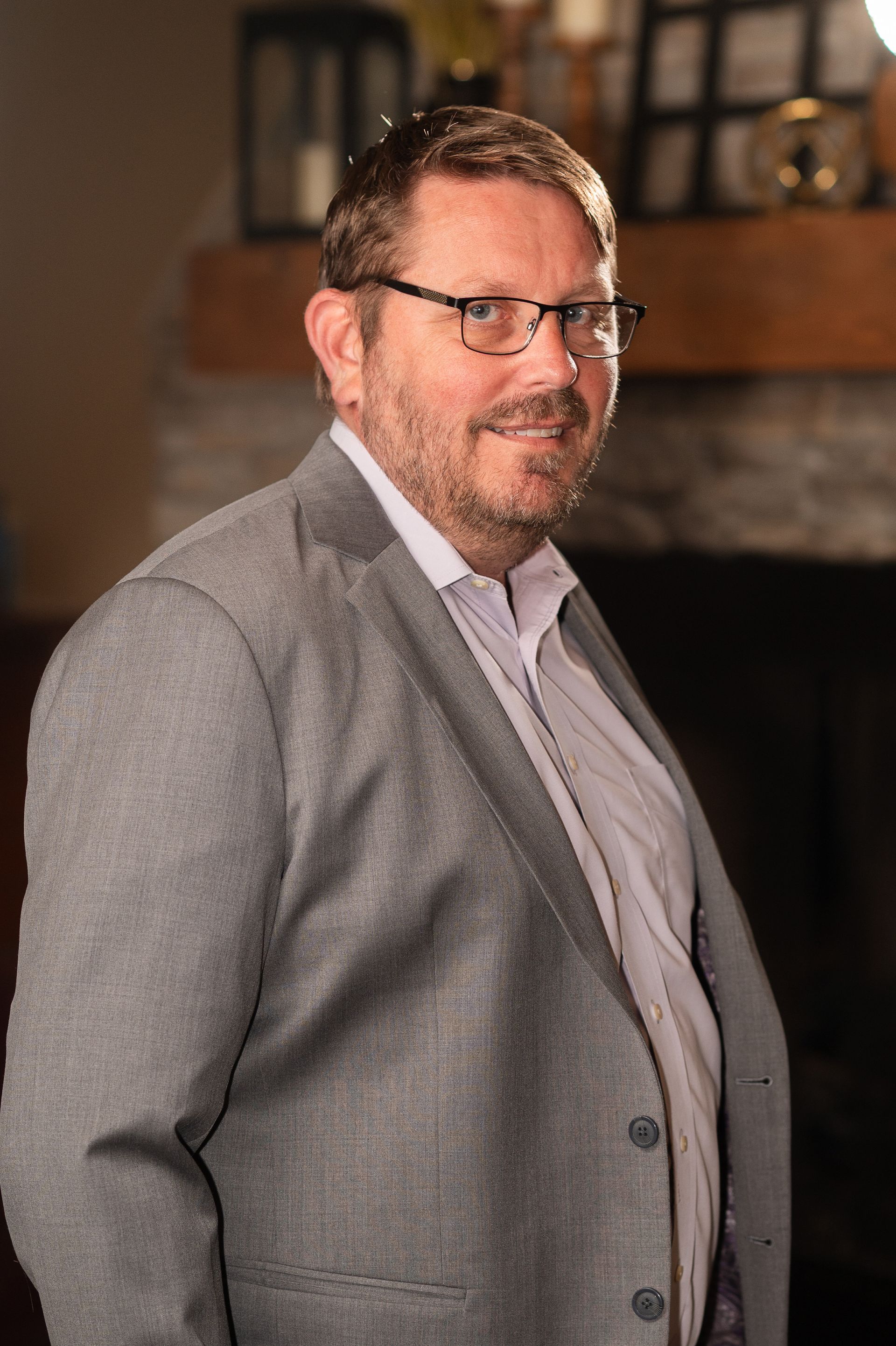A man in a suit and glasses is standing in front of a fireplace.