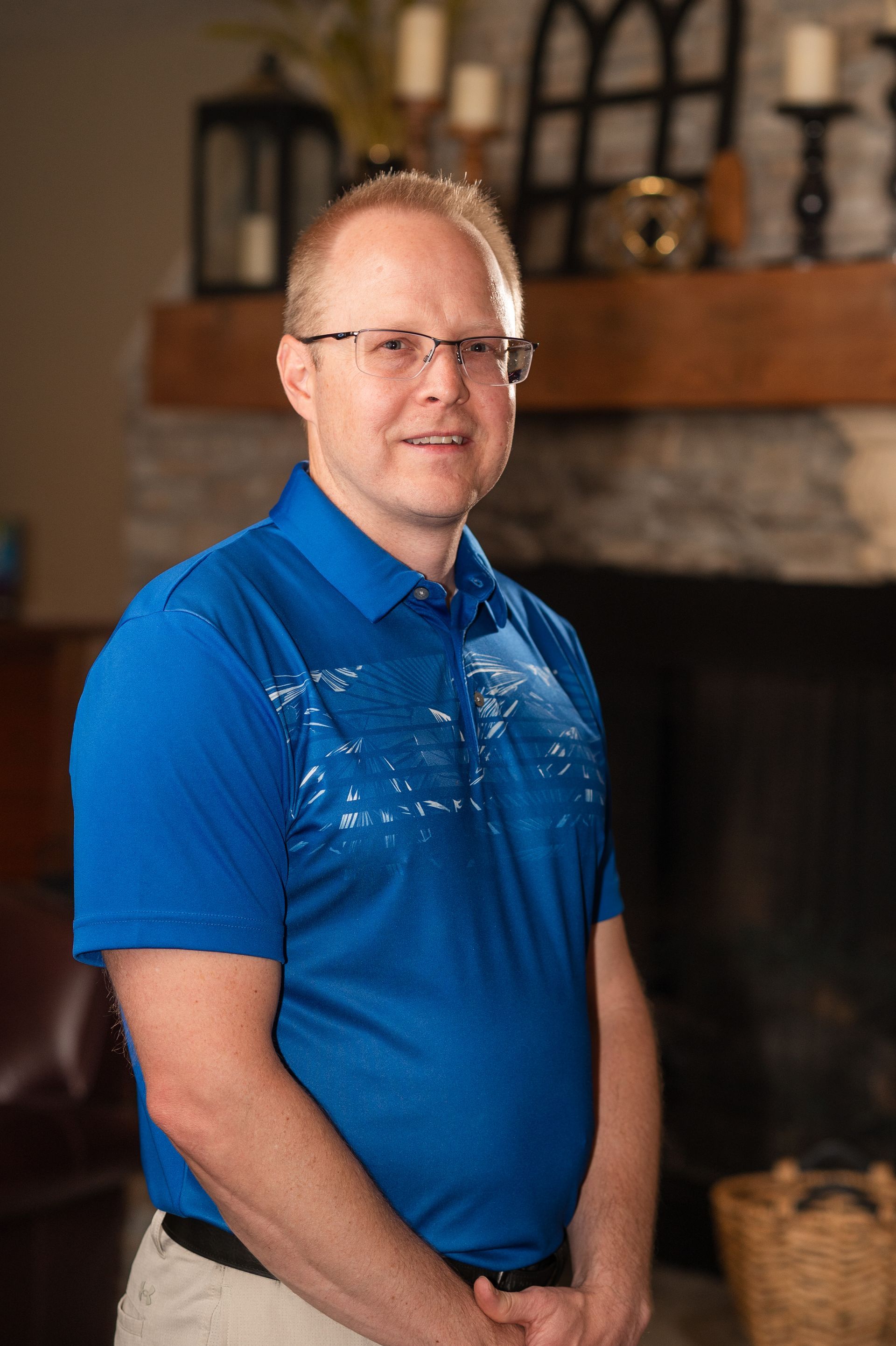 A man in a blue shirt and glasses is standing in front of a fireplace.