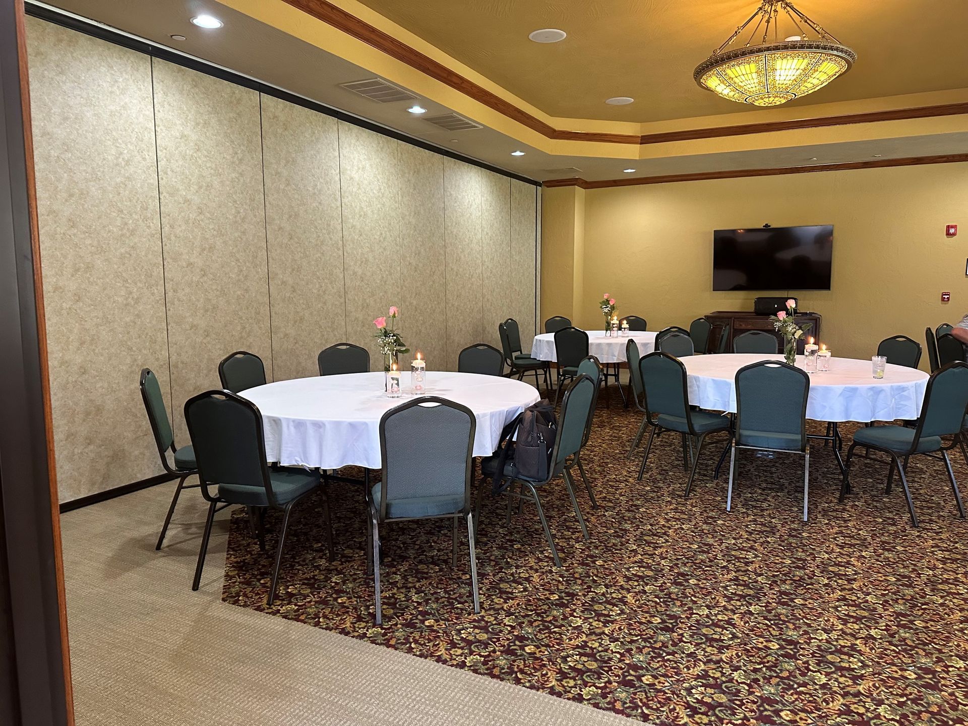 Banquet room with round tables, teal chairs, patterned carpet, and a wall-mounted TV.