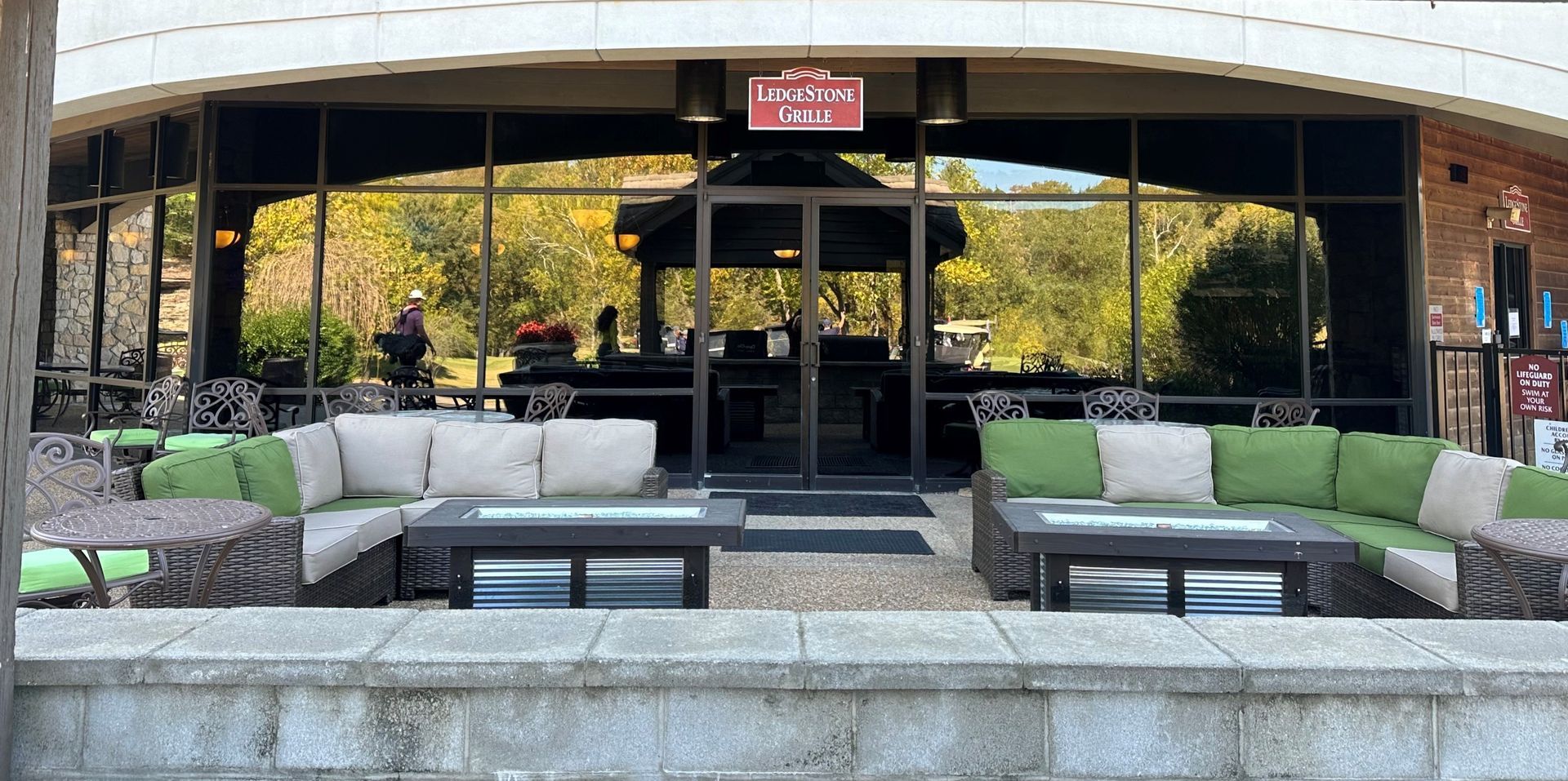 Covered outdoor lounge with gray sofas, green chairs, and glass doors under an awning