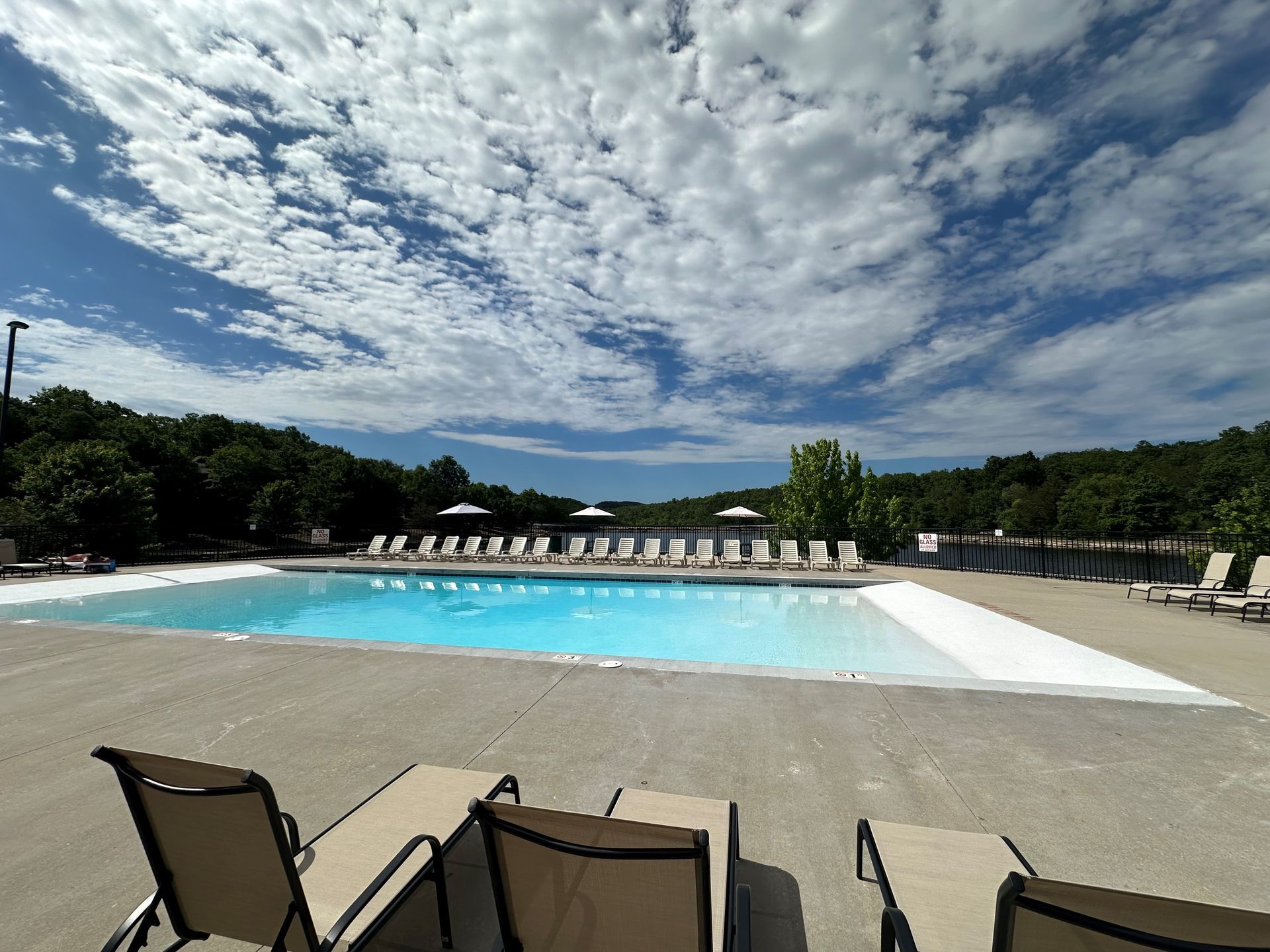 Outdoor swimming pool with lounge chairs under a cloudy sky, surrounded by trees and concrete deck