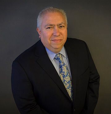 Man in dark suit and paisley tie smiles slightly against a gray background.