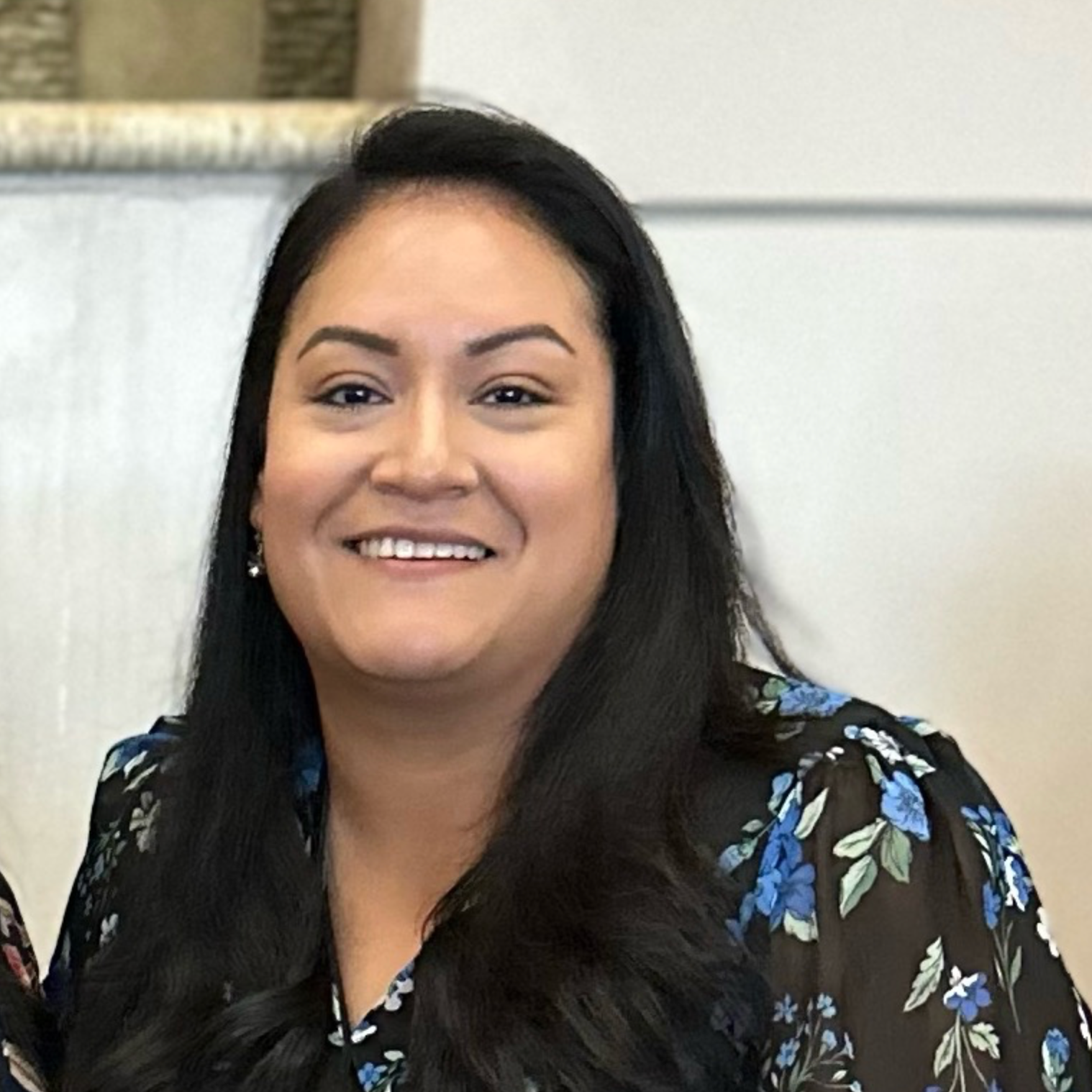 Woman with long dark hair smiles, wearing a floral-patterned top. Indoors, neutral background.