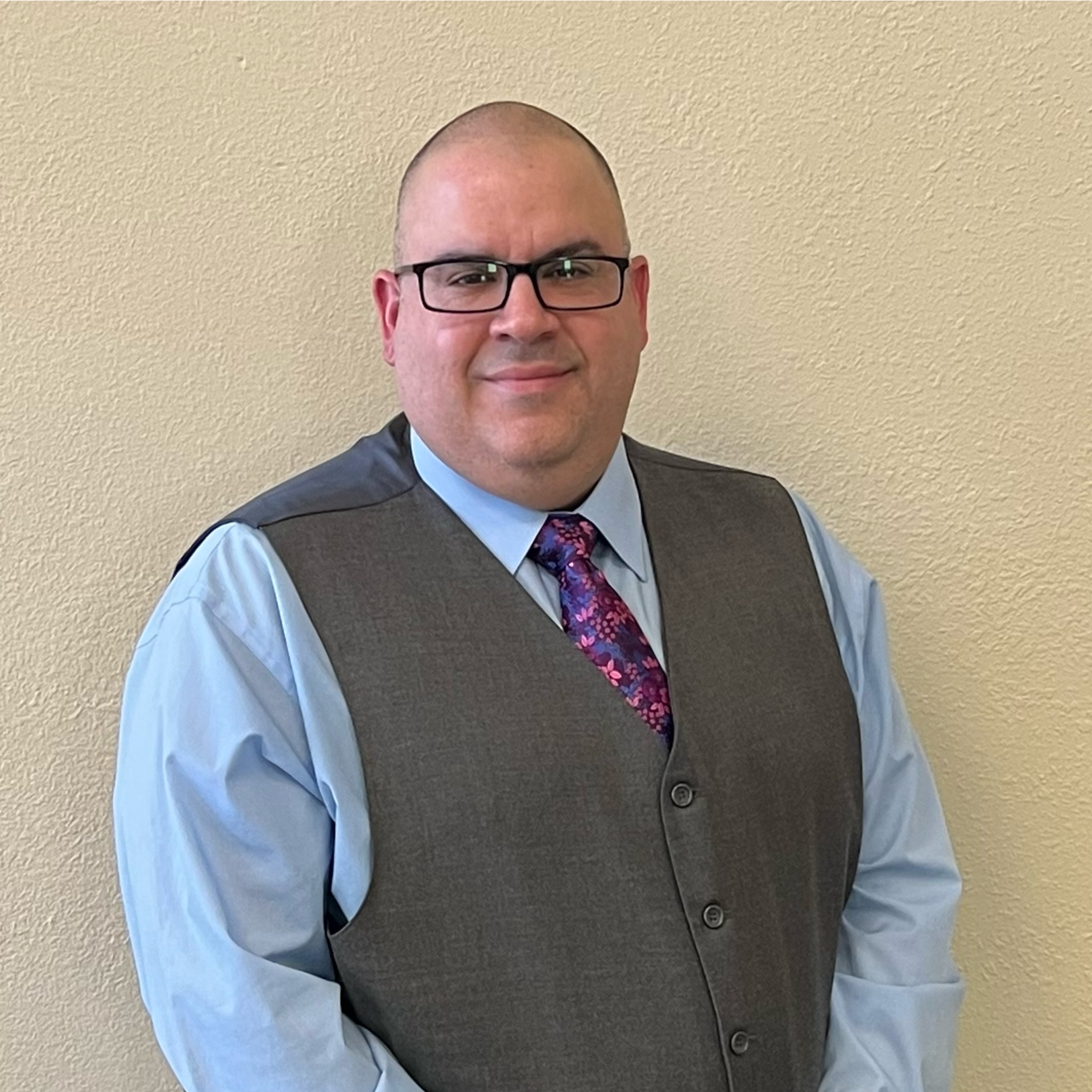 Man in glasses, grey vest, blue shirt, and patterned tie, posing in front of a tan wall.