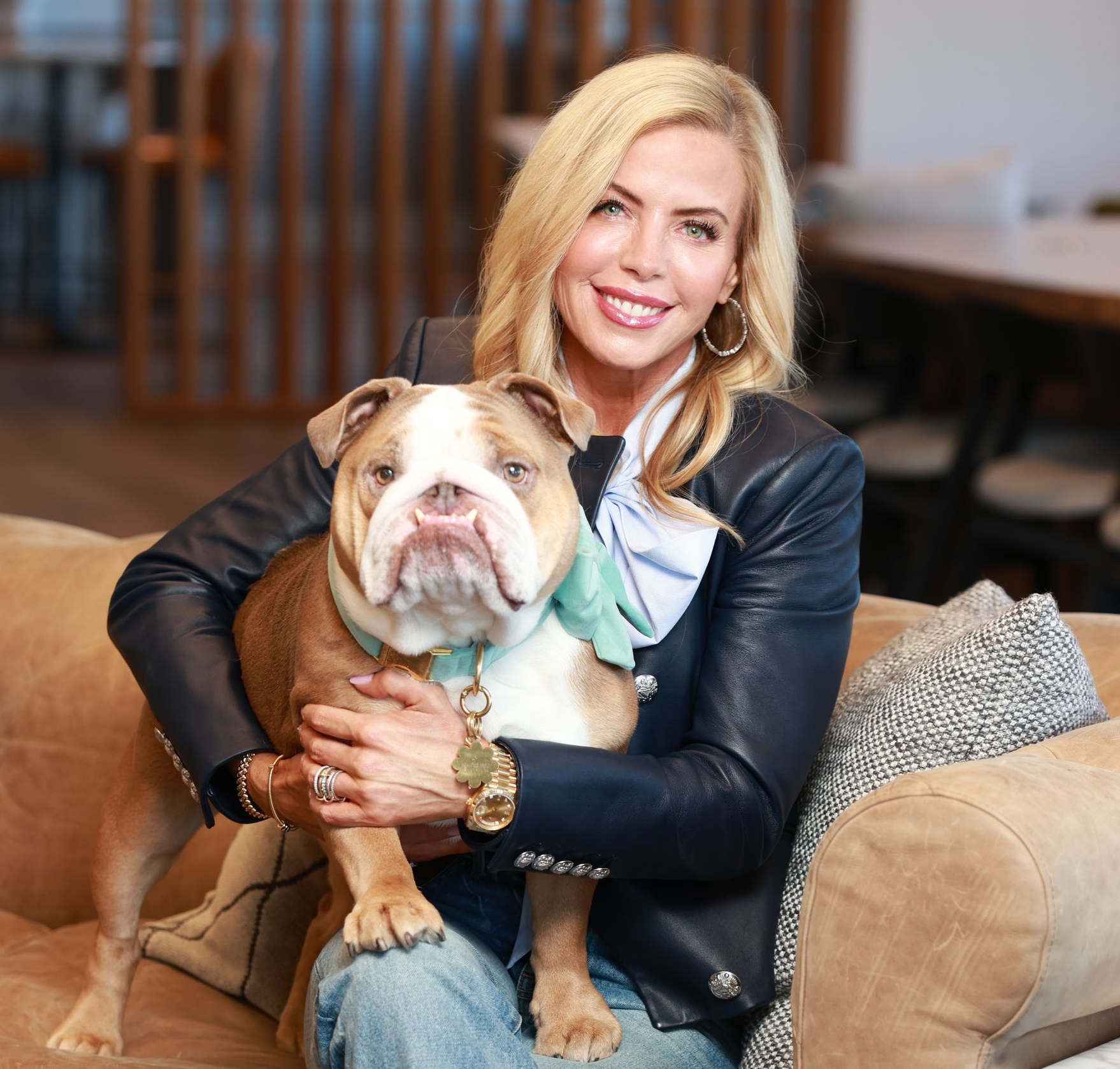 Blonde woman holding a bulldog on a couch in a cozy living room, both facing the camera.