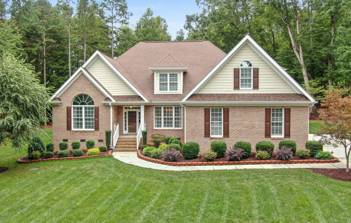 Brick house with manicured lawn, front porch, and landscaped garden surrounded by trees.