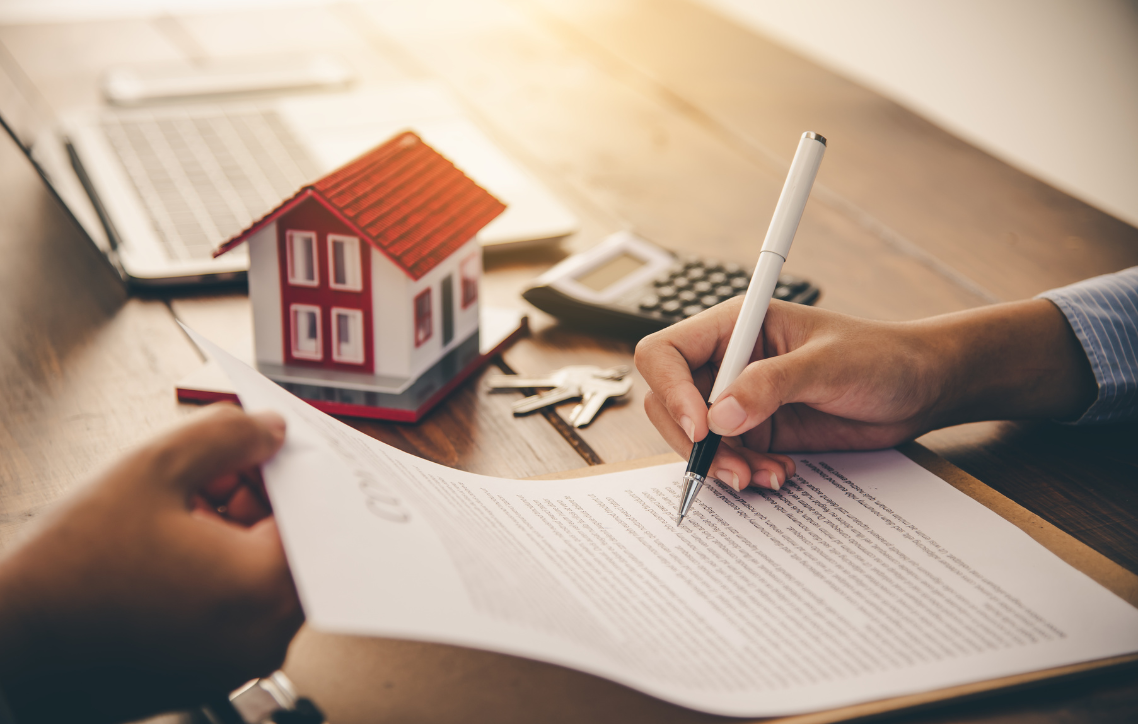 Hands signing a real estate contract beside a small house model and laptop on a desk