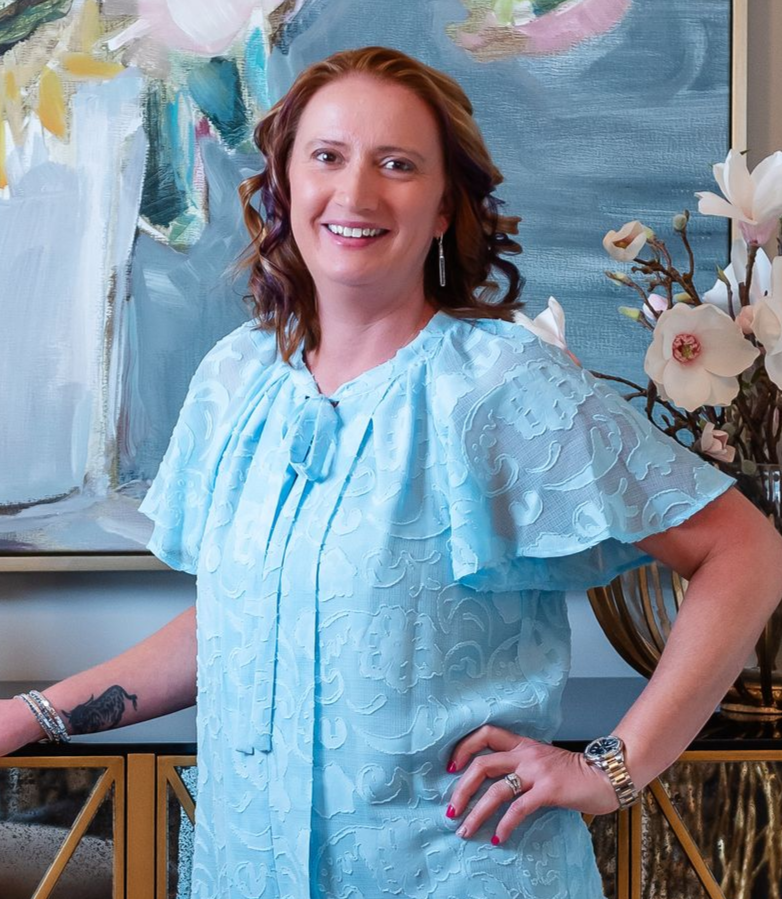 Smiling woman in a light blue blouse standing beside a gold console table and floral painting