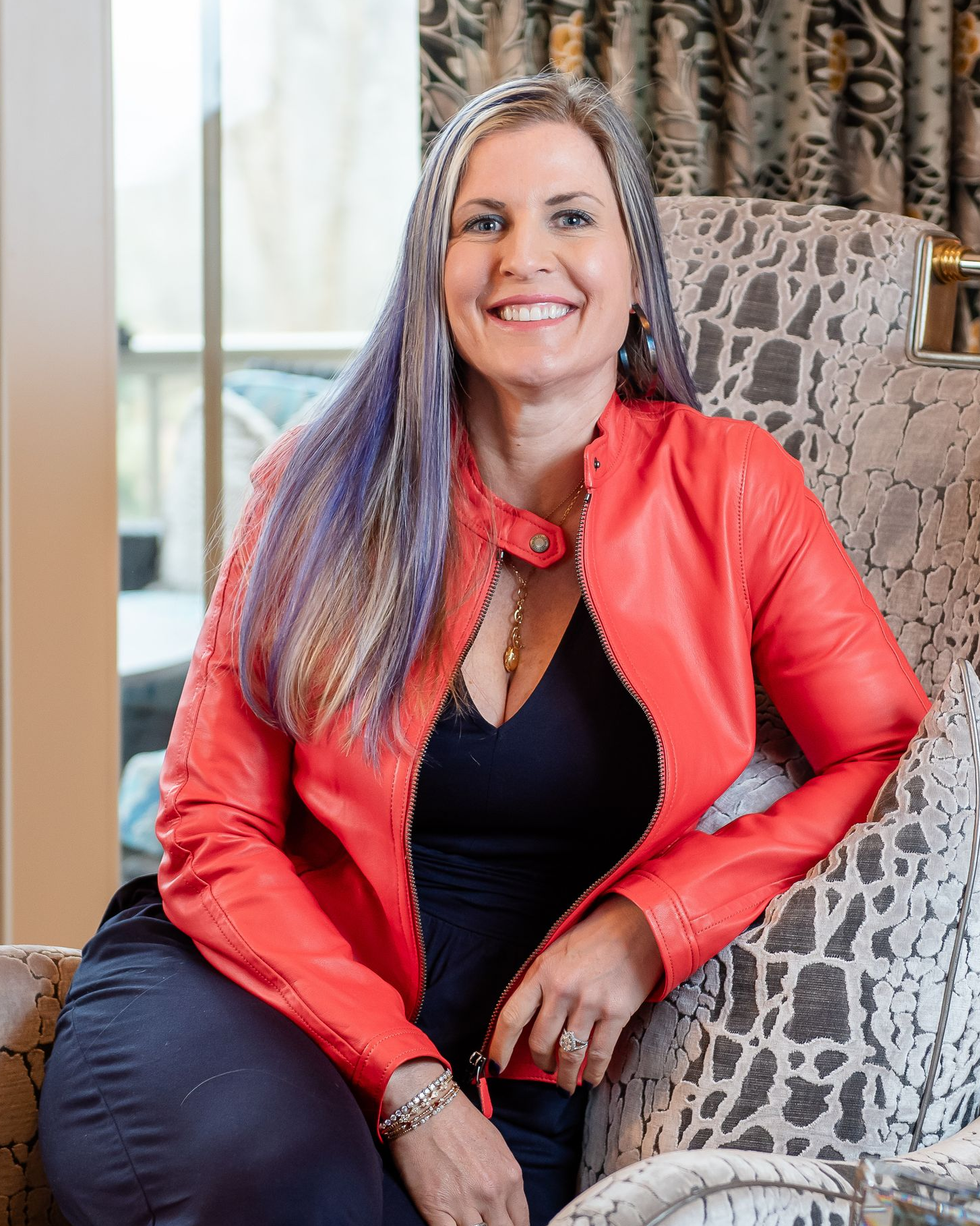 Woman in a red jacket smiling while seated in a patterned chair near a window.