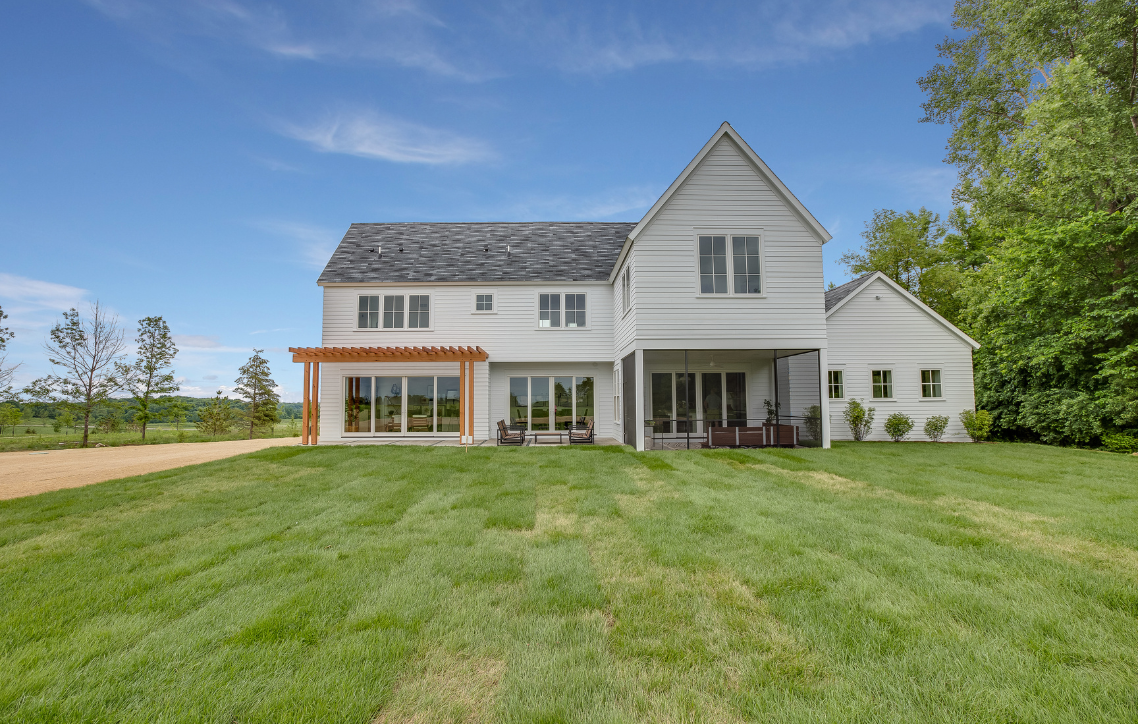 Large white farmhouse with a porch and attached barn on a grassy lawn under a blue sky
