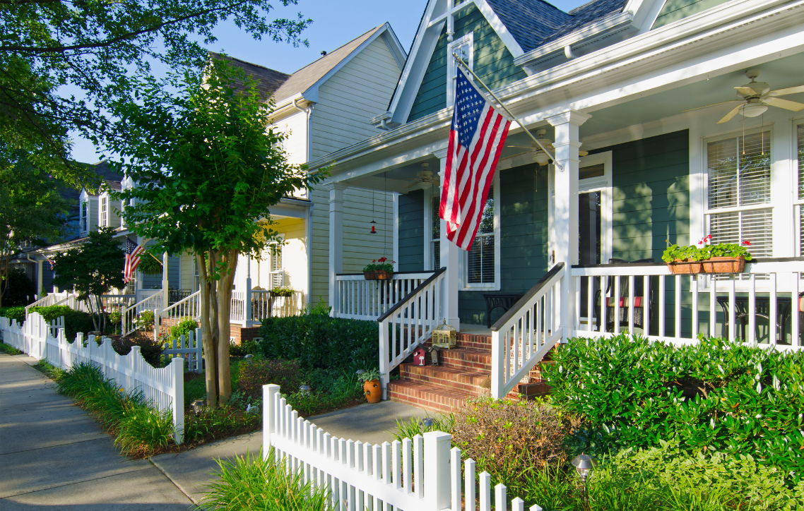 Row of white houses with porches and an American flag along a sunny tree-lined sidewalk