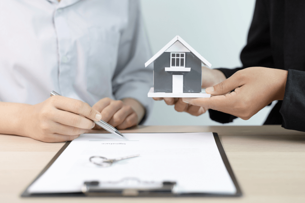 A person is holding a model house while another person signs a document.