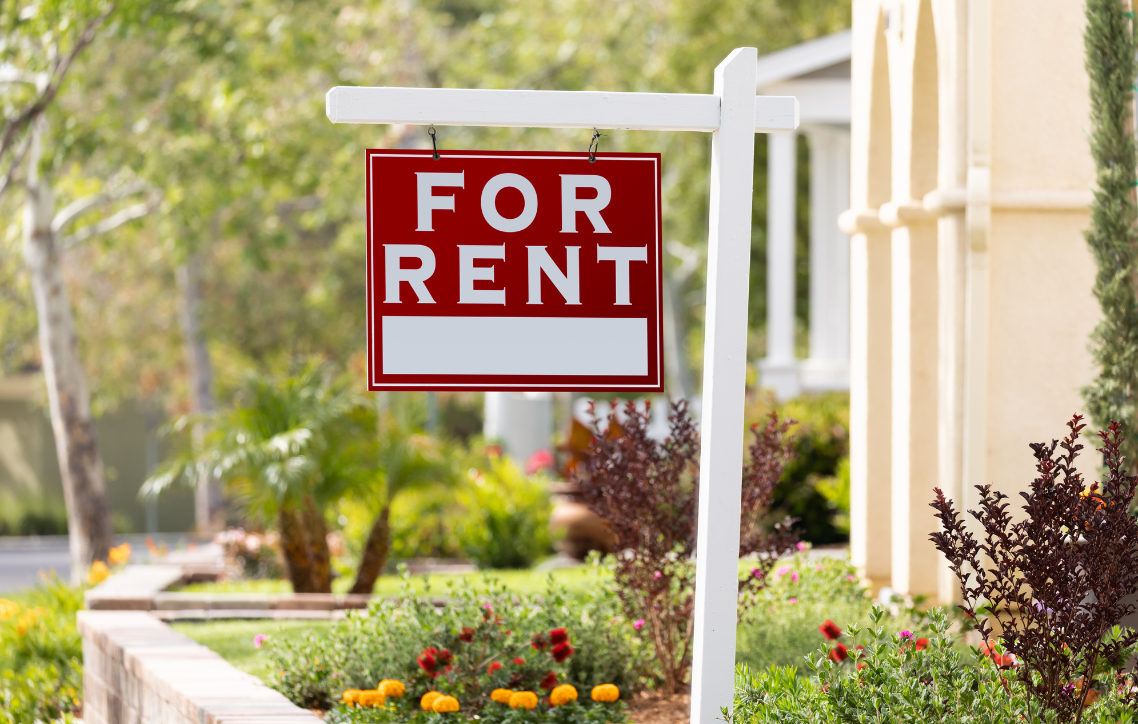 Red “For Rent” sign beside a white fence in a sunny garden yard