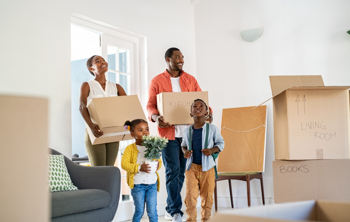 Family carrying moving boxes in a bright living room, suggesting they are unpacking in a new home