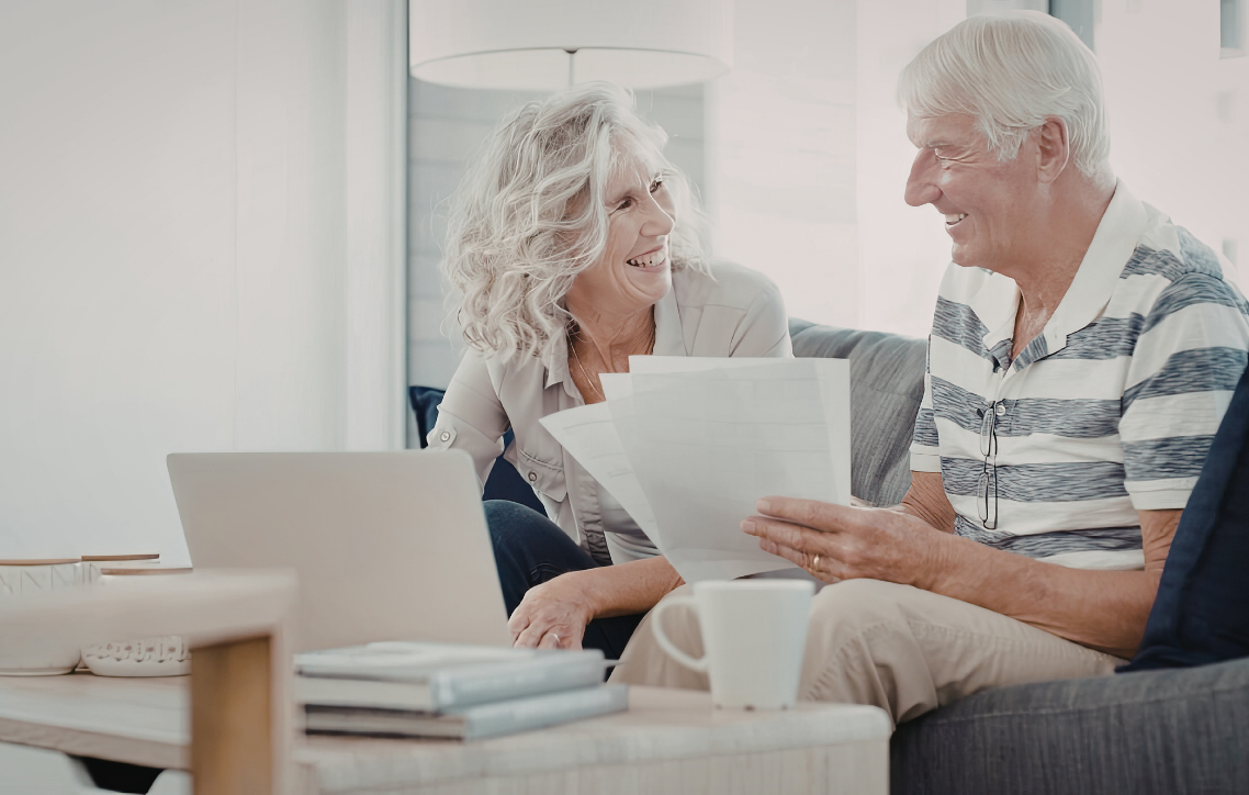 Two smiling seniors sit on a couch, reviewing papers in a bright living room.