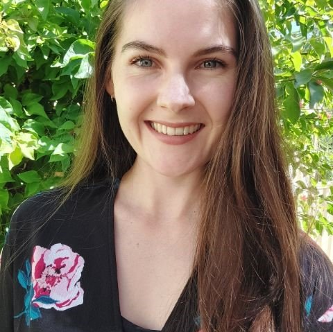 Woman with long brown hair smiles, wearing a black floral robe, in front of greenery.