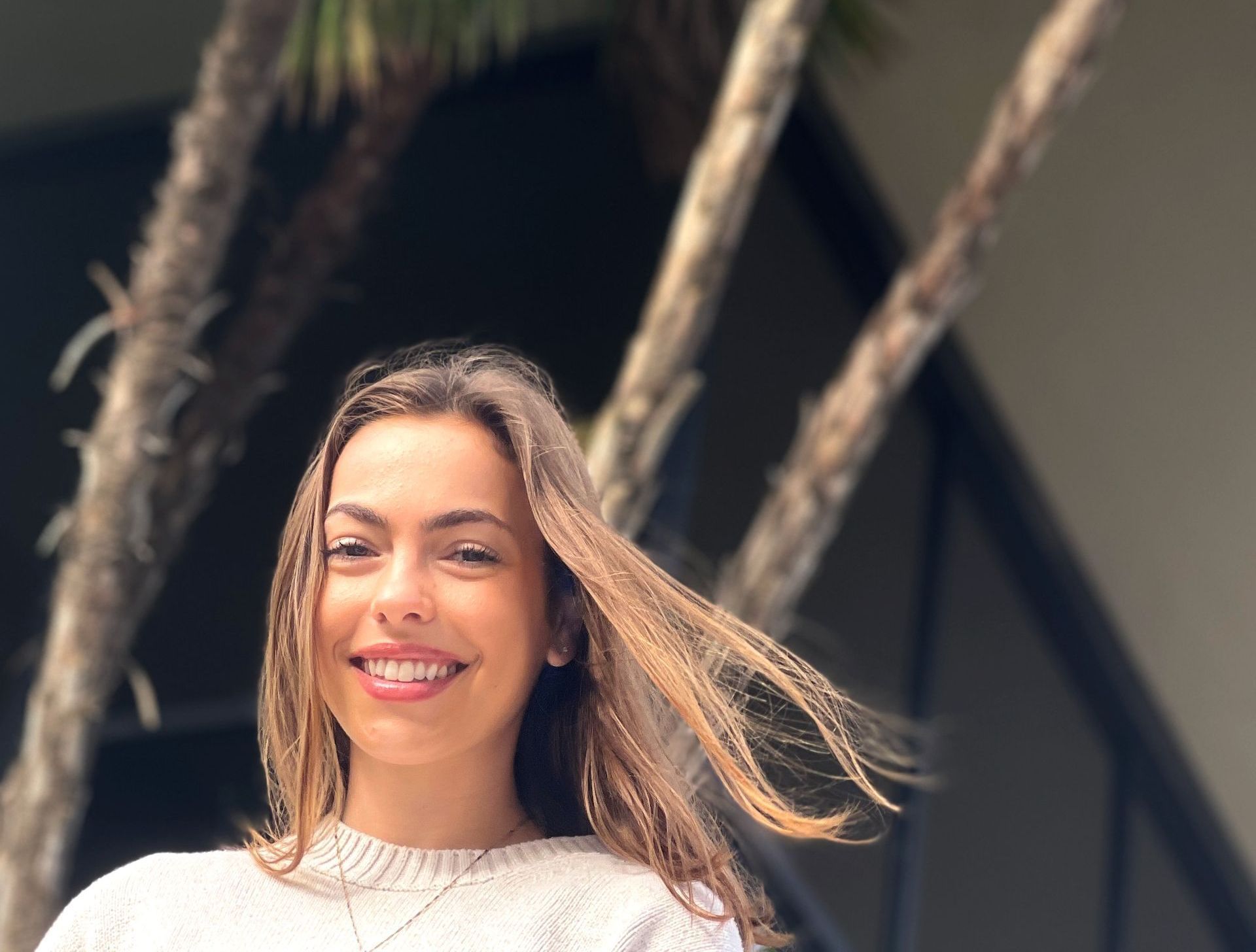 A woman in a white sweater is smiling in front of a palm tree