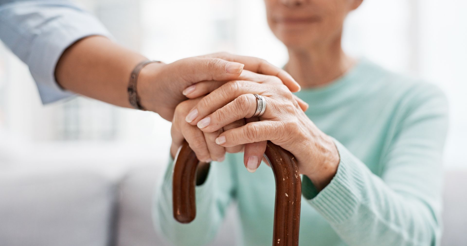 A person's hand rests on an older woman's hand, both over a walking cane.