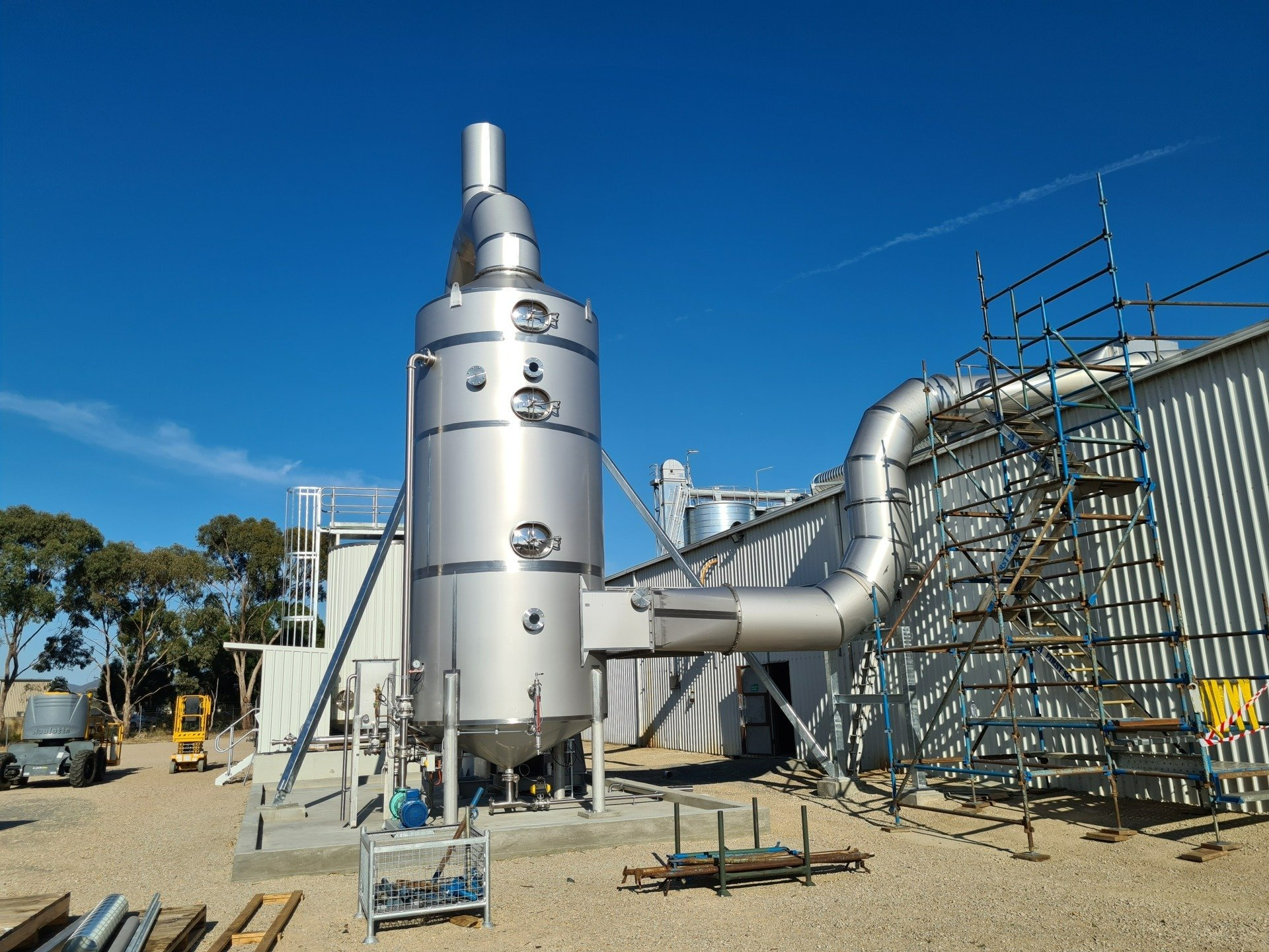 Industrial stainless steel scrubber system outdoors. Metal structure with a curved exhaust pipe against a bright blue sky.