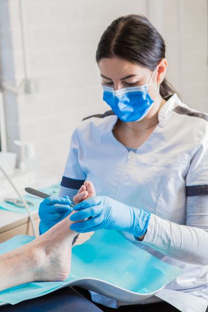 Een vrouw draagt ​​een masker en handschoenen terwijl ze de teennagels van een patiënt knipt.