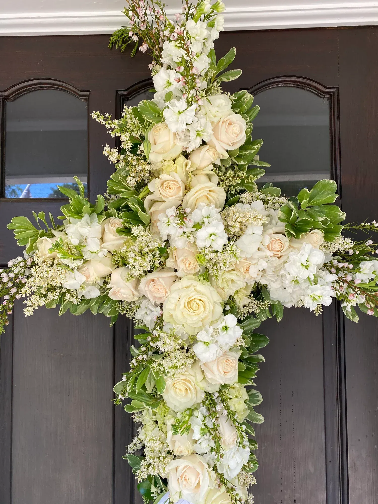 Floral cross made of white roses and greenery, hanging on a brown door.