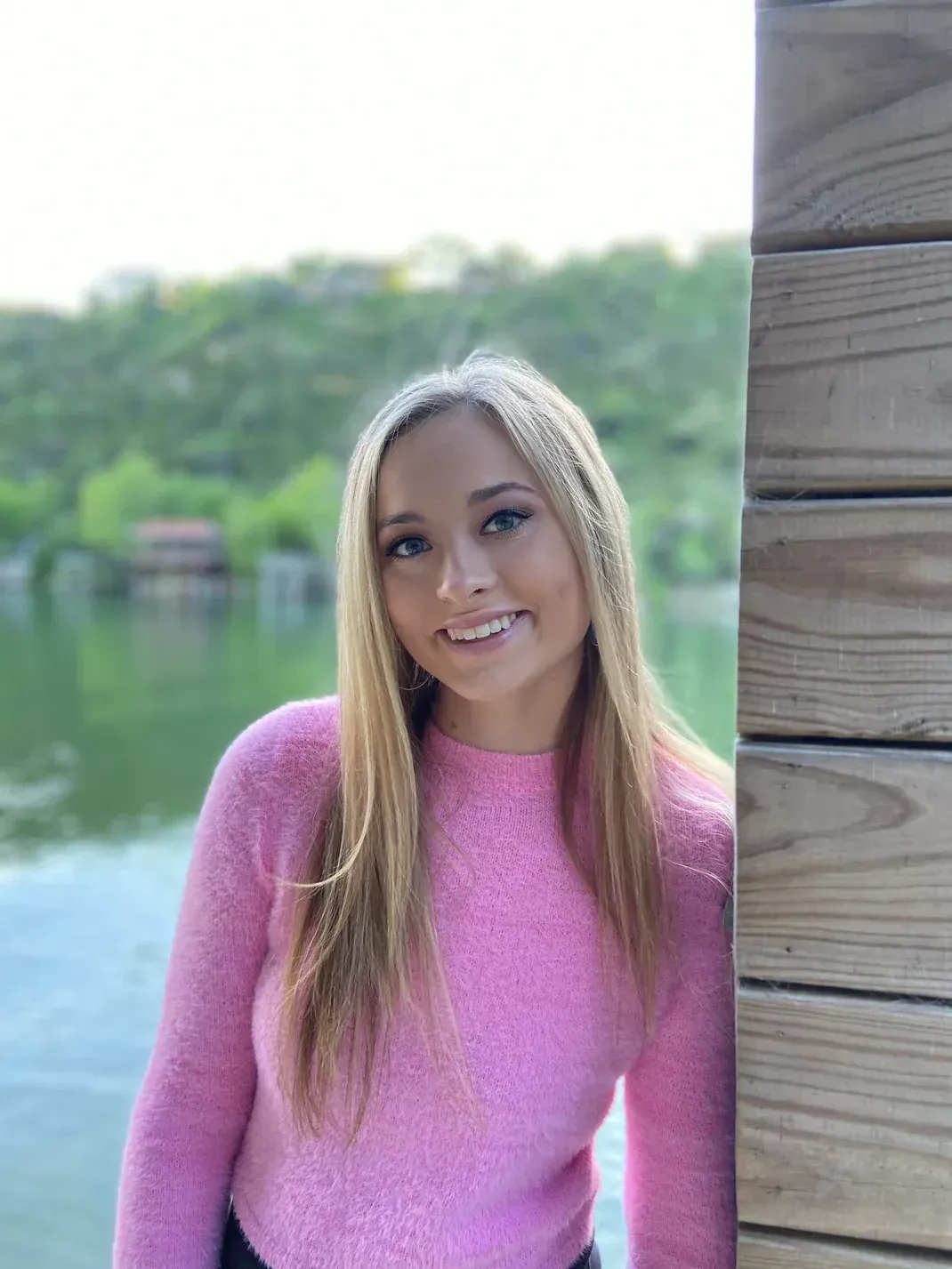 Blonde person in pink sweater smiles, leaning on wooden structure by lake.