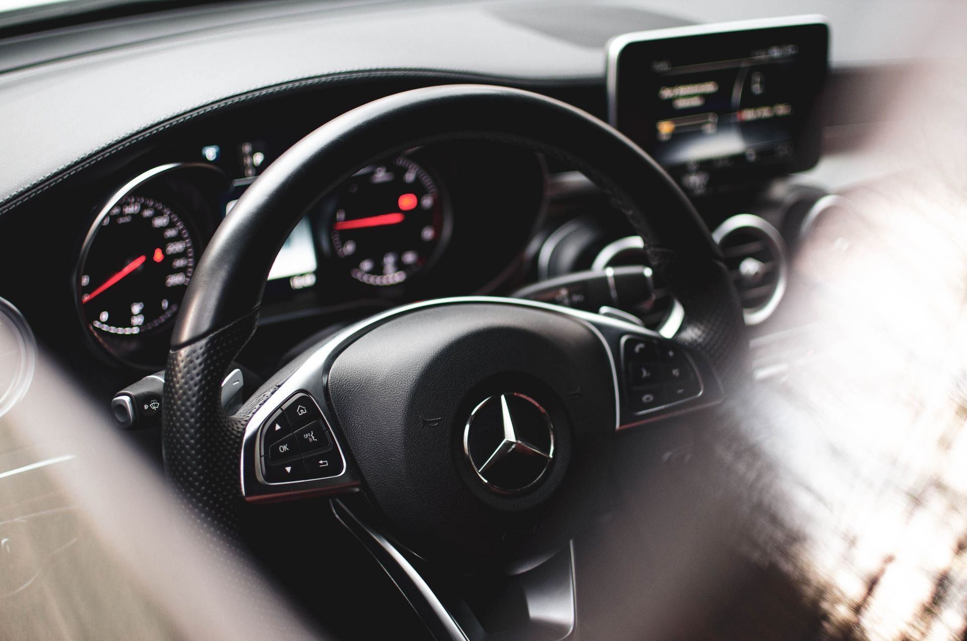 Black Mercedes steering wheel with controls, dashboard, and logo in the center.