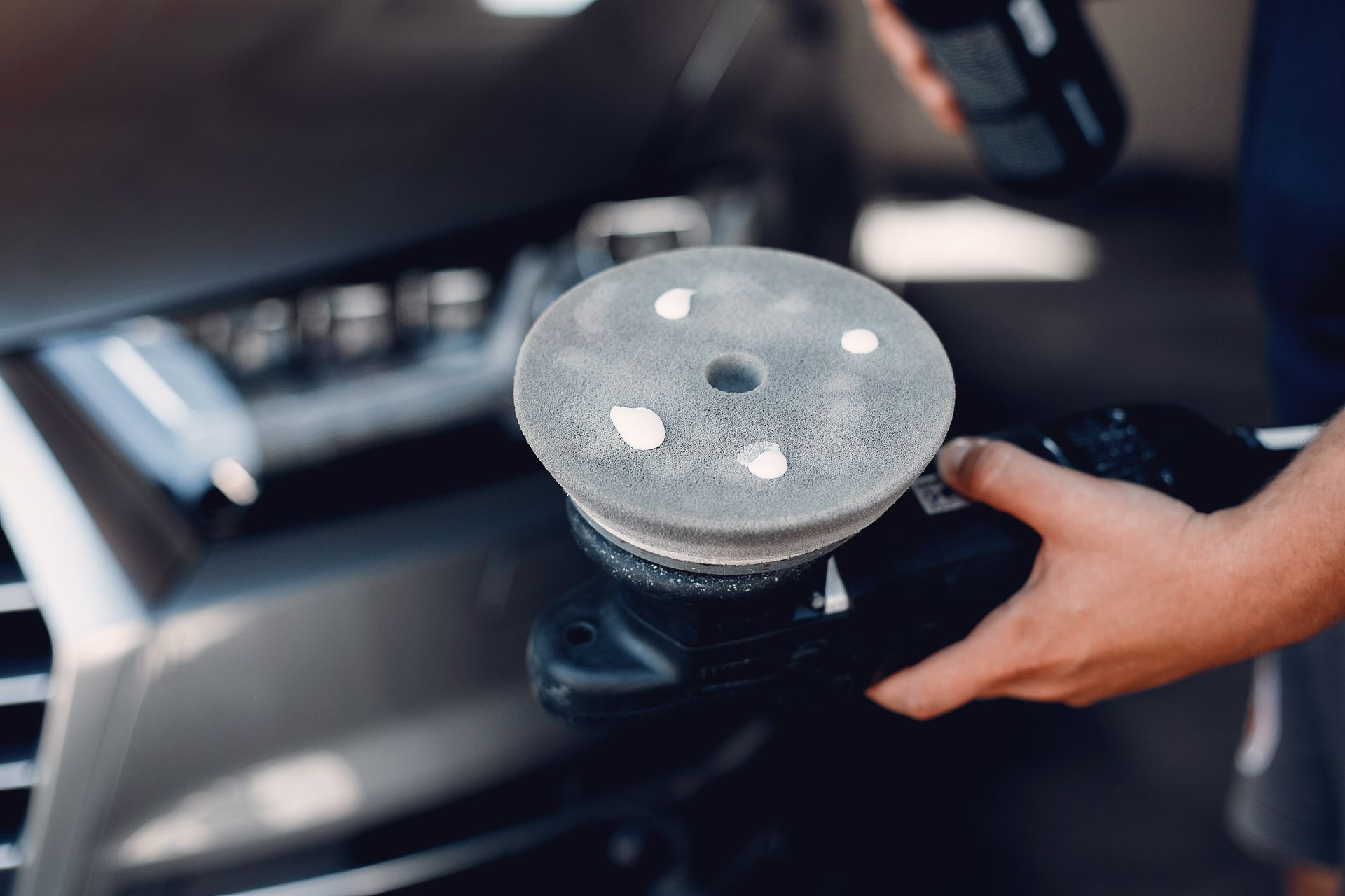 Person polishing a car's dark hood with a polishing machine, holding a spray bottle.