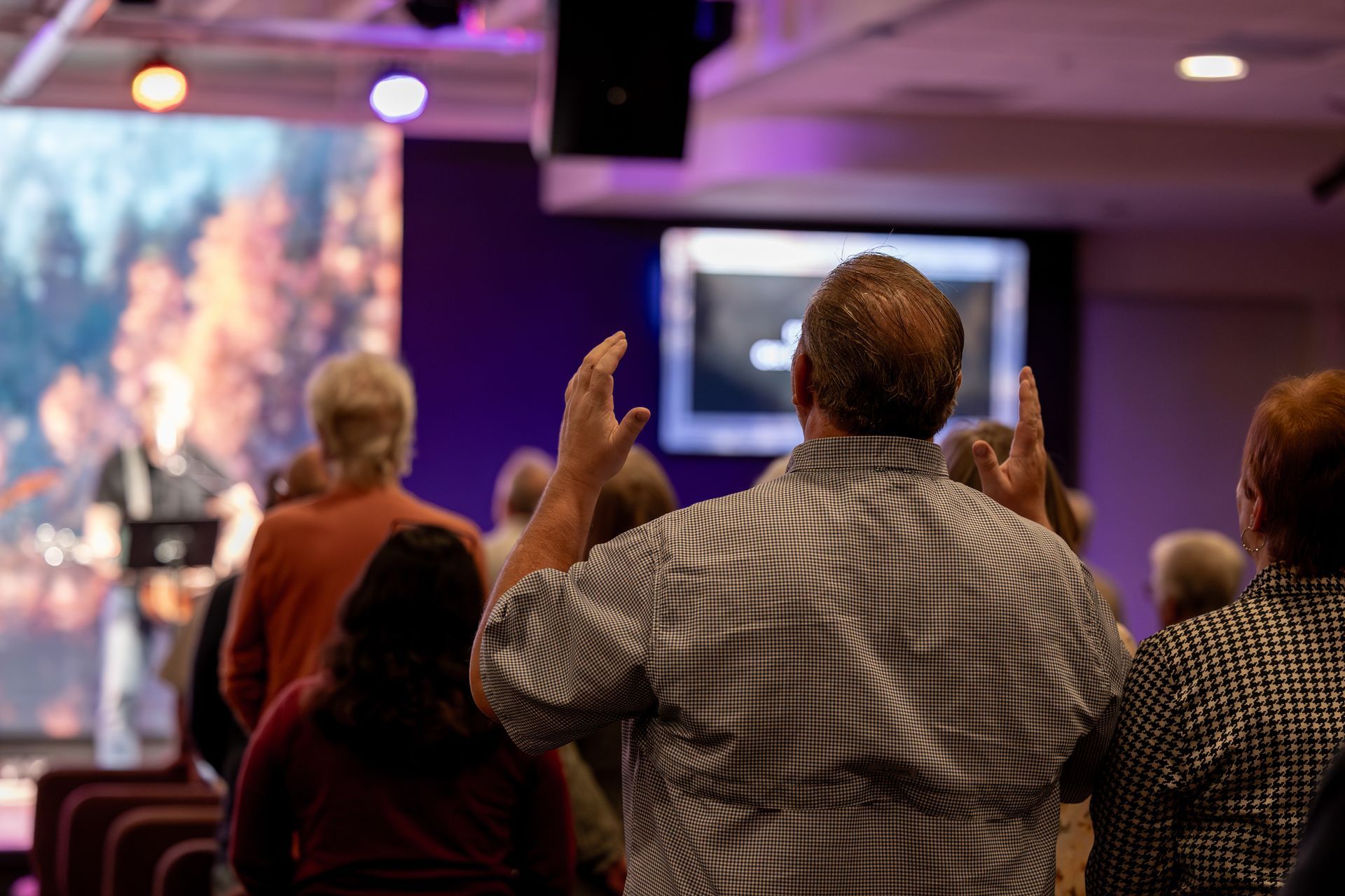 Audience standing and raising hands during a presentation in a dim conference room with screens and purple lighting